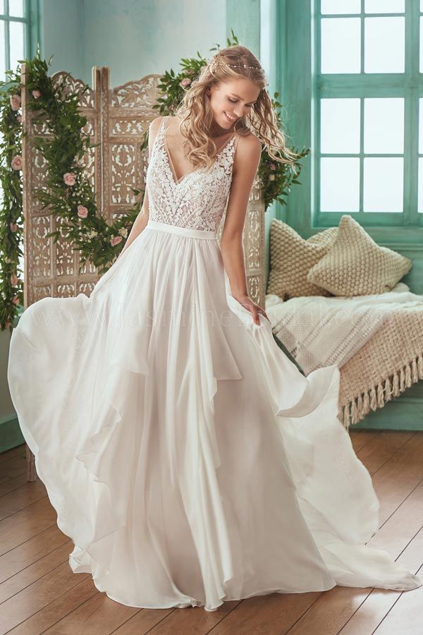 Woman in a white lace wedding dress standing in a bright room beside a bed and window.