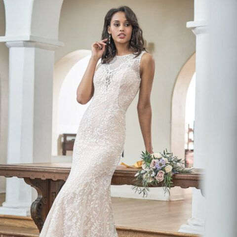 Woman in a white lace gown posing indoors beside a wooden table with flowers and white columns
