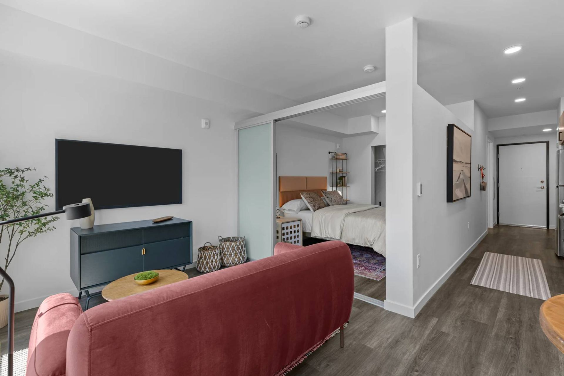 Open living area with red sofa, wall-mounted TV, and sliding door to a bedroom at Zinnia Apartments in Shoreline, WA.