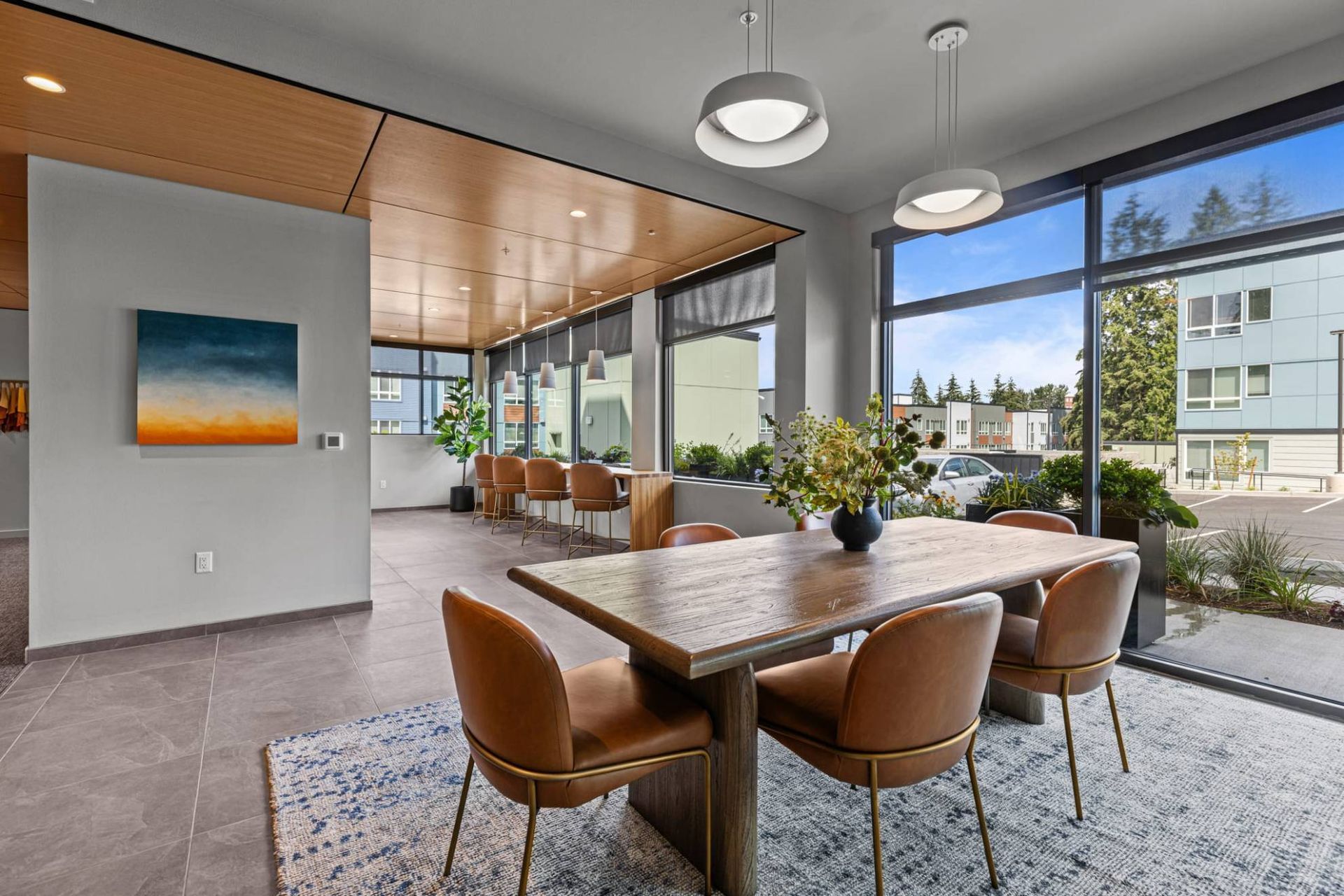 Bright communal lounge with large windows, a wooden dining table, and tan chairs at Zinnia Apartments in Shoreline, WA.