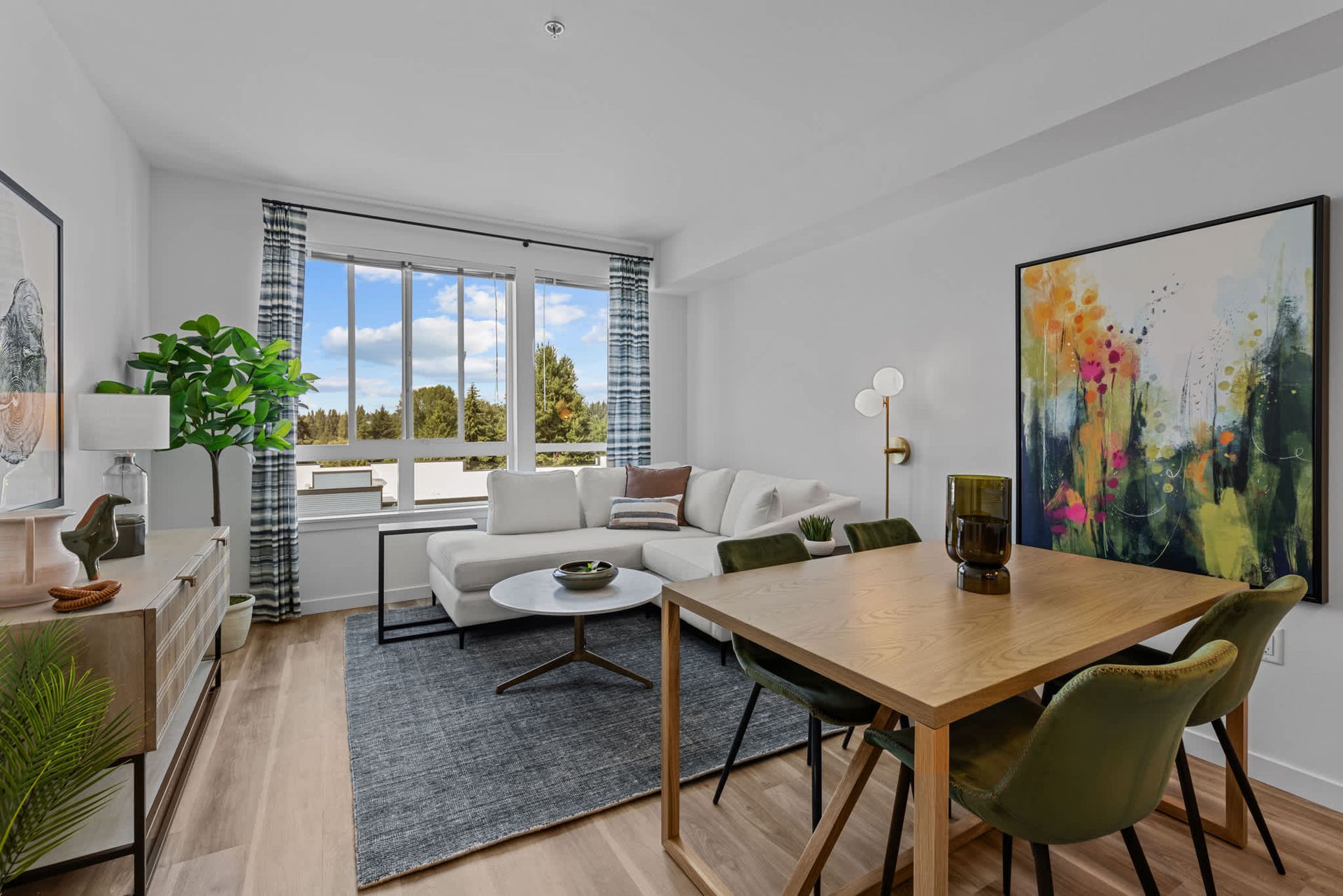 Bright living room in a modern apartment with a white sectional, wooden dining table, and large window at Zinnia Apartments in Shoreline, WA.