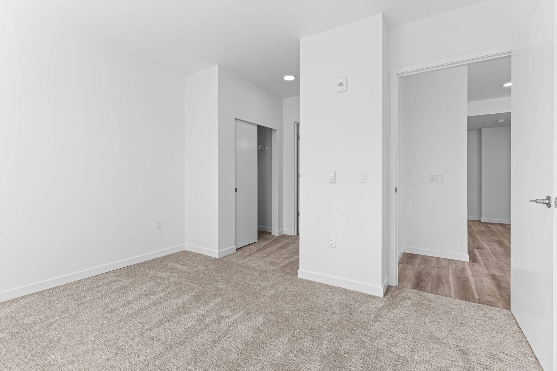 Empty bedroom with beige carpet, white walls, and a closet; doorway to a room with wood flooring at Zinnia Apartments in Shoreline, WA.