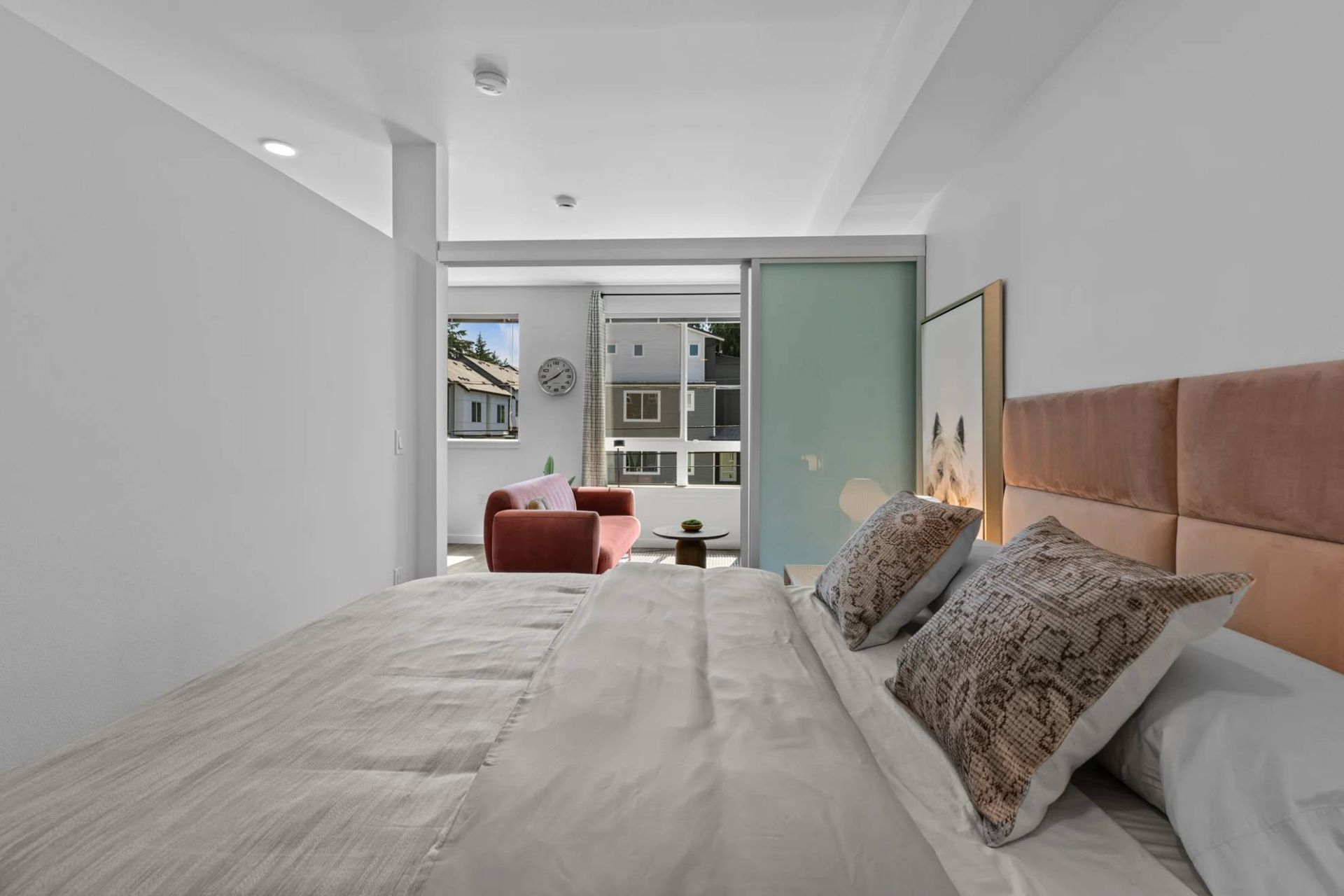 Bedroom with white bedding and padded headboard, sliding glass door to living area at Zinnia Apartments in Shoreline, WA.