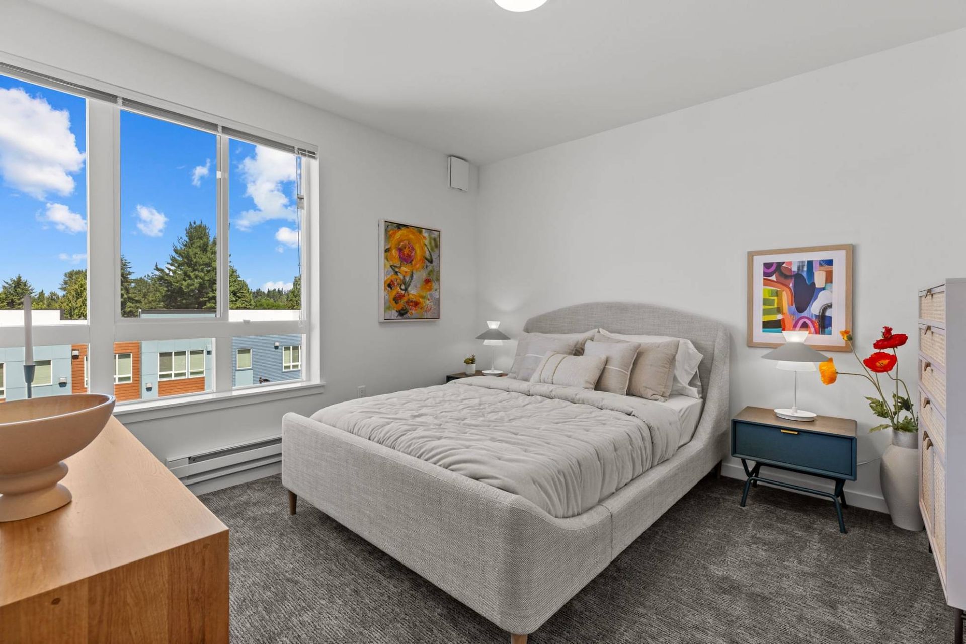 Bright bedroom in a modern apartment with a gray upholstered bed and a large window at Zinnia Apartments in Shoreline, WA.