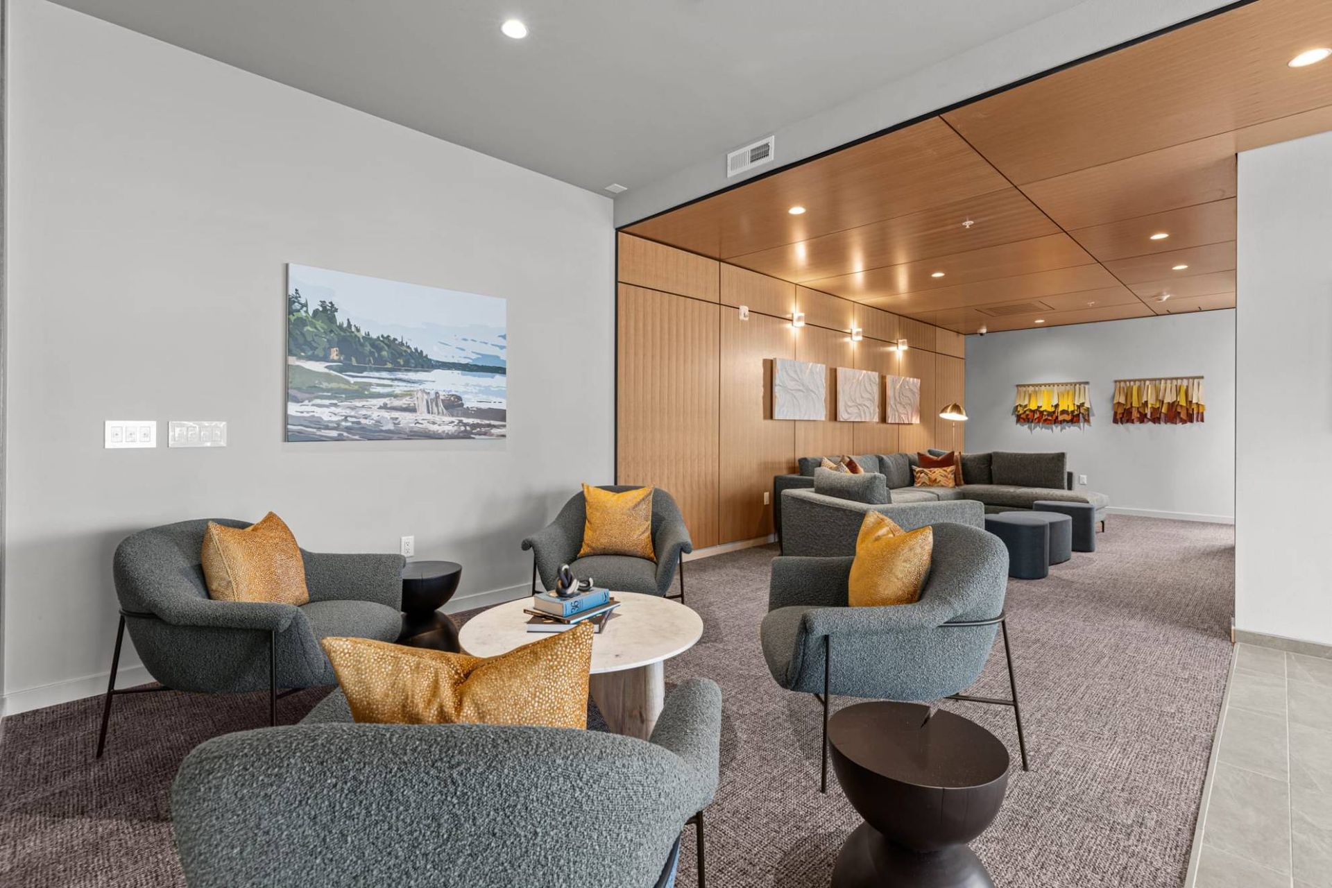 Modern apartment community lounge with grey seating, orange pillows, wood paneling, and wall art at Zinnia Apartments in Shoreline, WA.