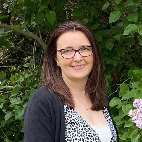 A woman wearing glasses and a leopard print shirt is standing in front of a bush.