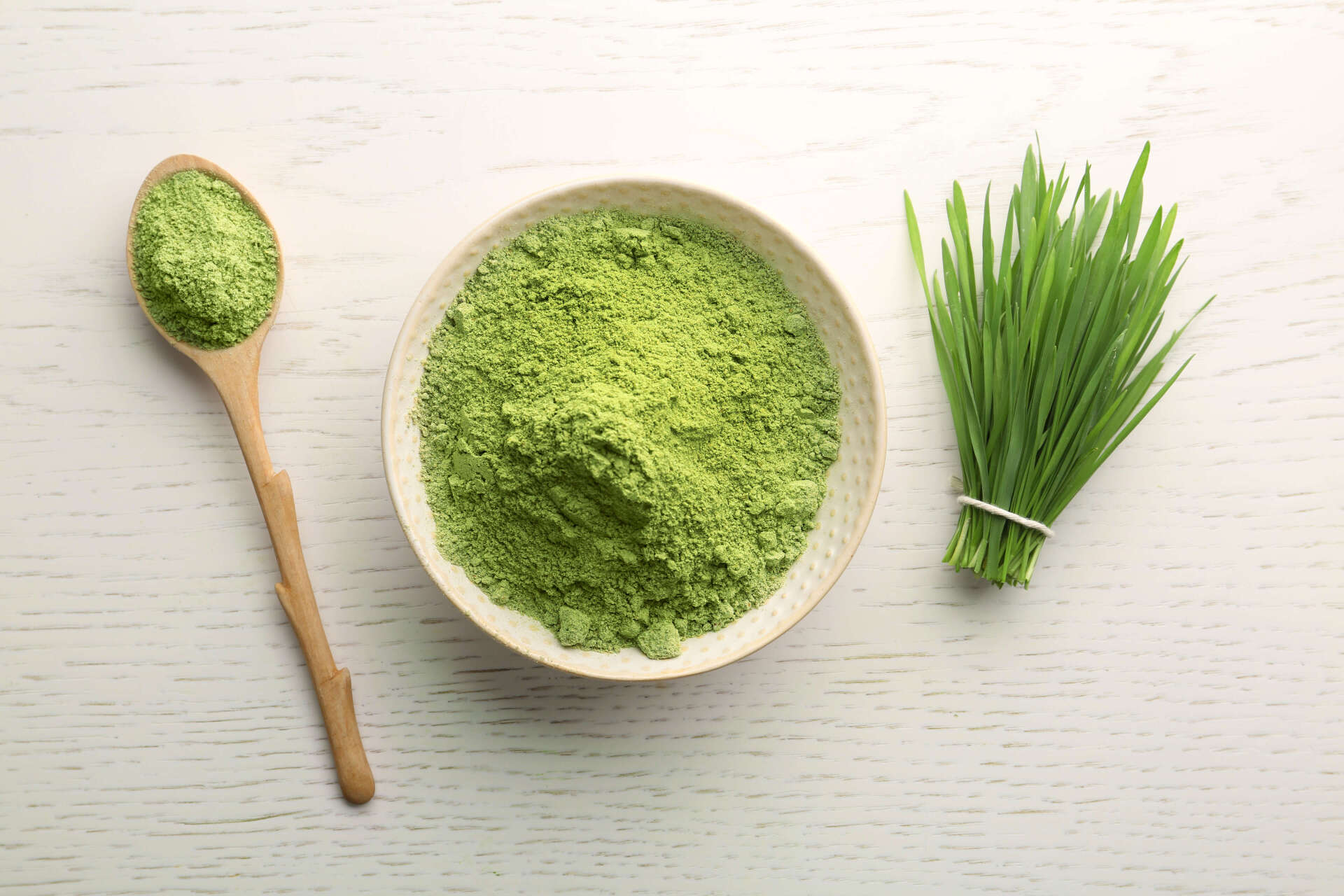 A bowl of wheat grass powder next to a wooden spoon and a bunch of wheat grass.