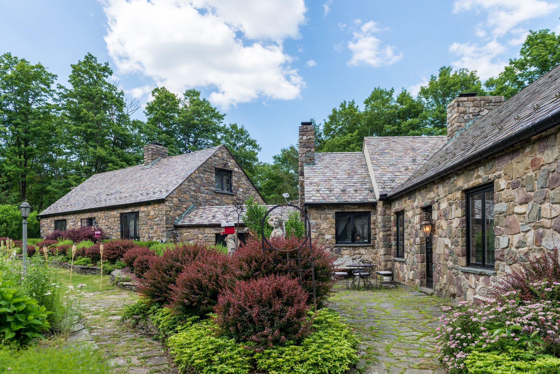 A stone house with a slate roof is surrounded by bushes and trees.