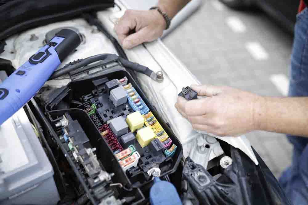 a close up of a fuse box under the hood of a car with a mechanic hands holding a fuse