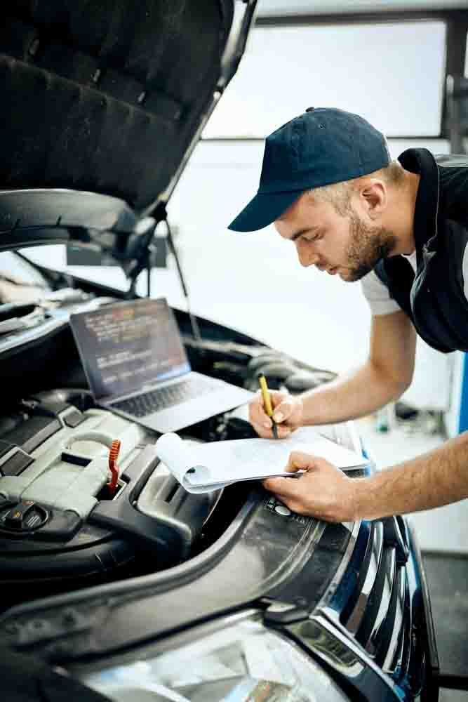a mechanic in a blue hat looking under the hood of a car with a laptop and a clipboard performing a vehicle diagnostic