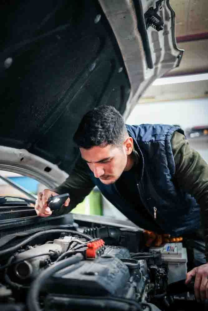 A mechanic looking under the hood of a car at the engine bay with a flashlight