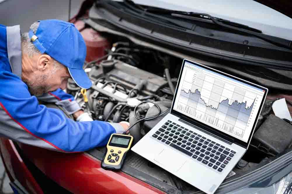 mechanic in a blue hat performing vehicle diagnostic in the engine bay with a laptop