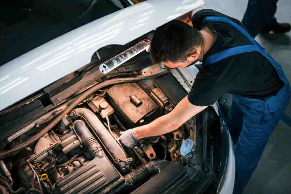 mechanic in blue overalls looking at the engine bay of a car with a flashlight