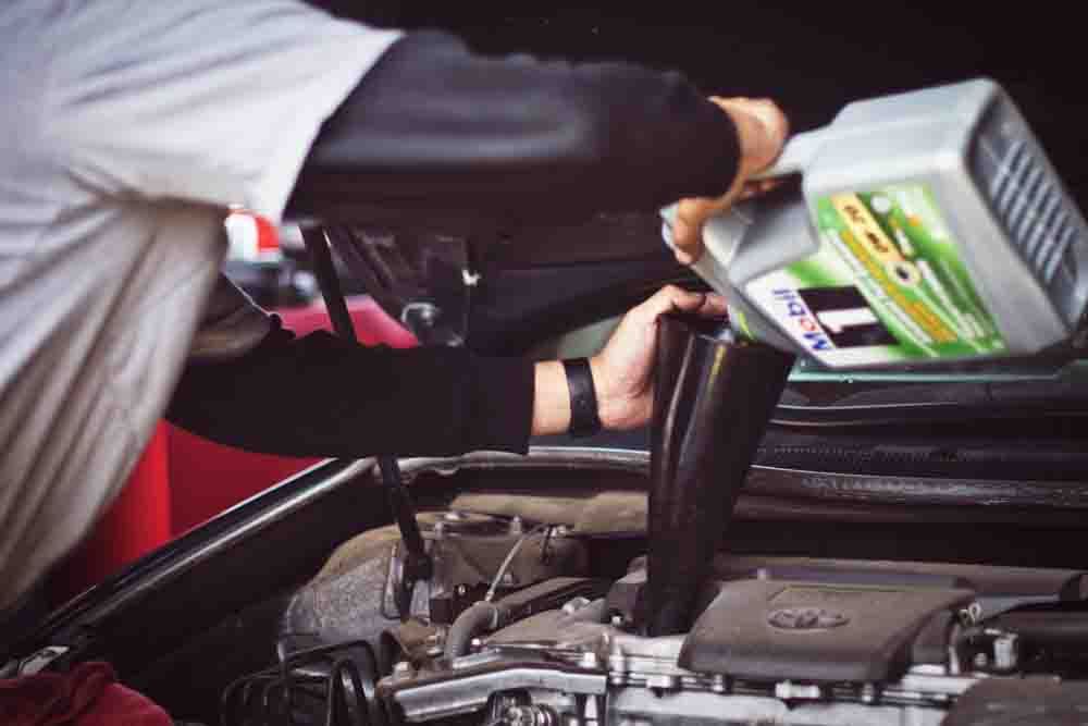 a mechanic in a grey shirt pouring fresh new oil into a funnel and a car engine