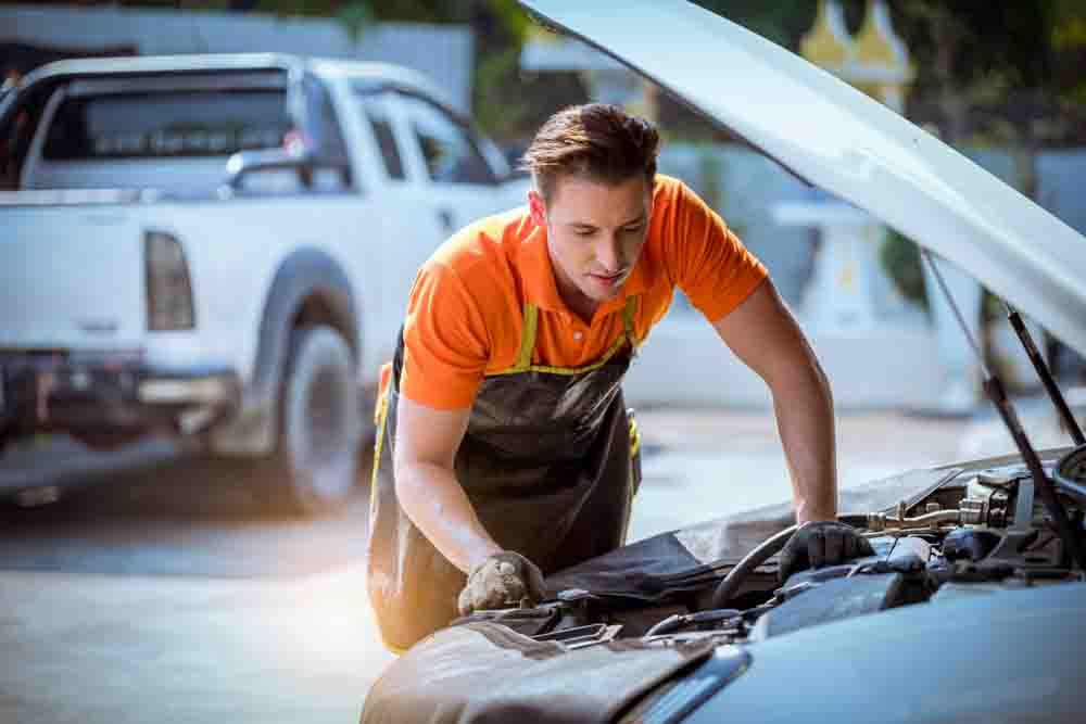 A mechanic wearing an orange shirt with an apron looking under the hood of a car