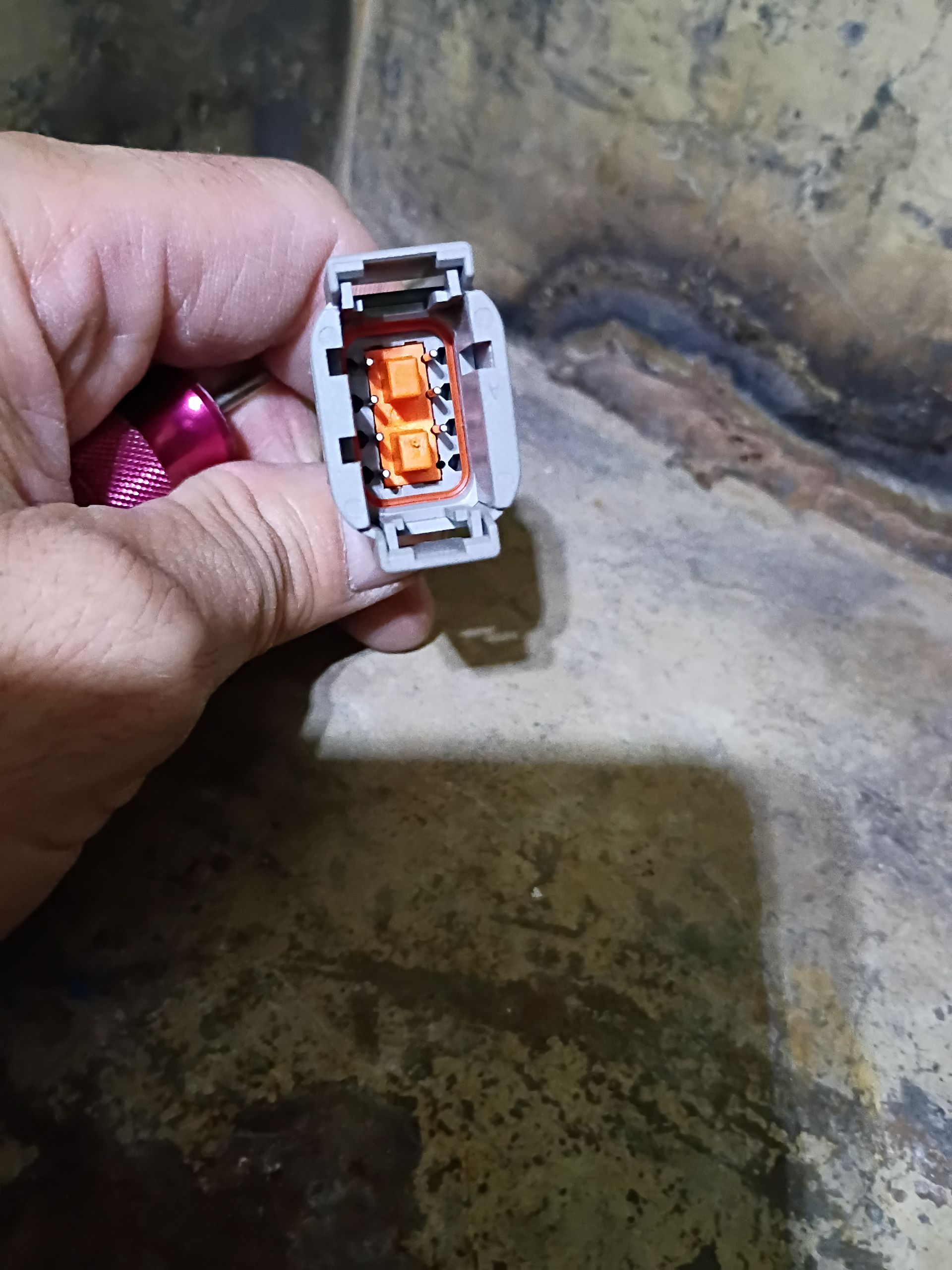 Hand holding a white electrical connector with orange insert over a dirty concrete floor
