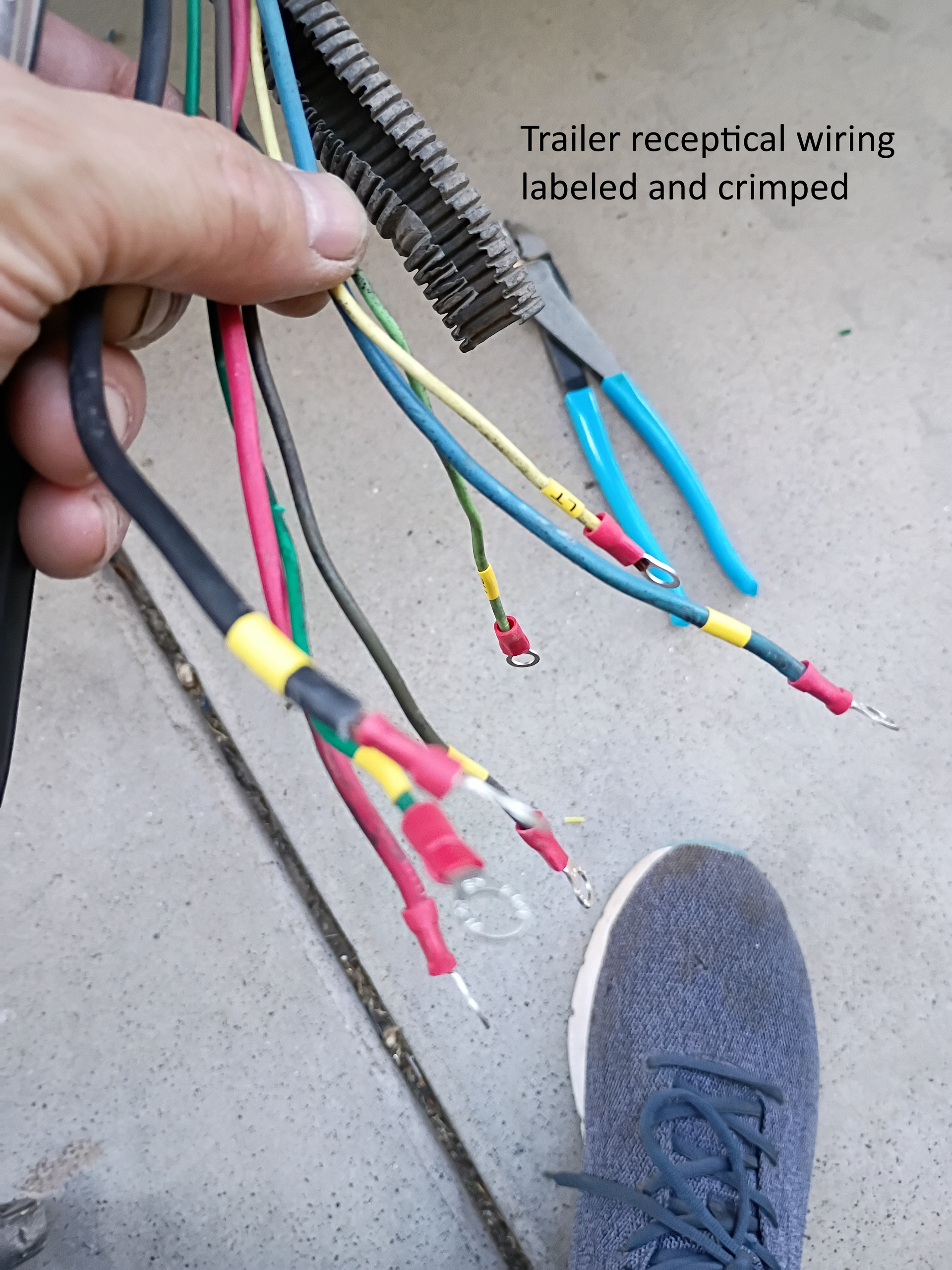 Hand holding labeled trailer receiver wiring with colored wires and crimped connectors on concrete beside a shoe