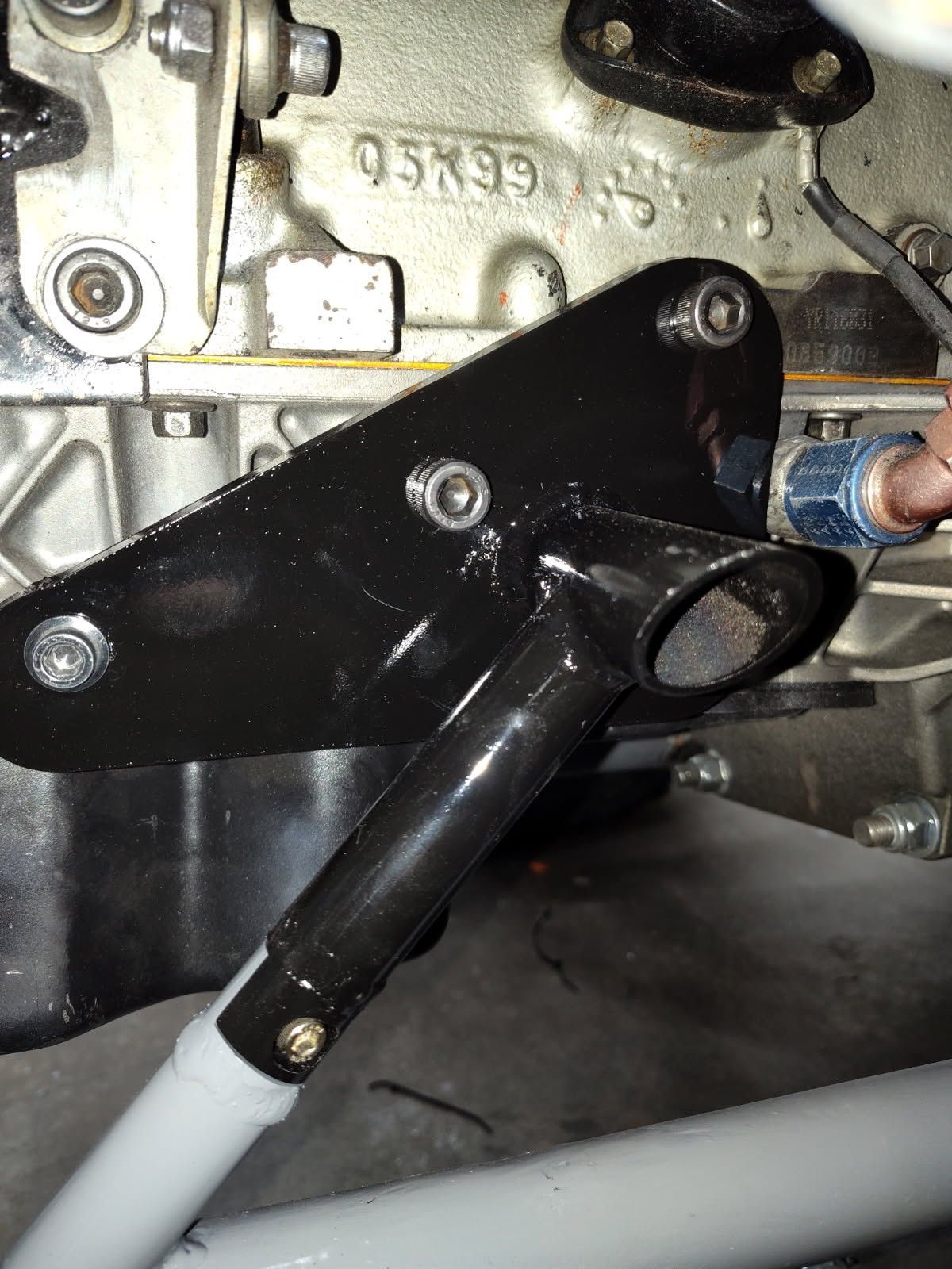 A black metal bracket mounted to an engine block, with a gray tubular support arm extending downward.