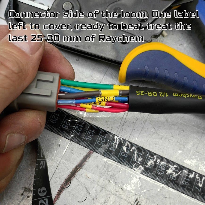 Hand holding a gray connector with multicolored wires and yellow heat-shrink tubing on a workbench