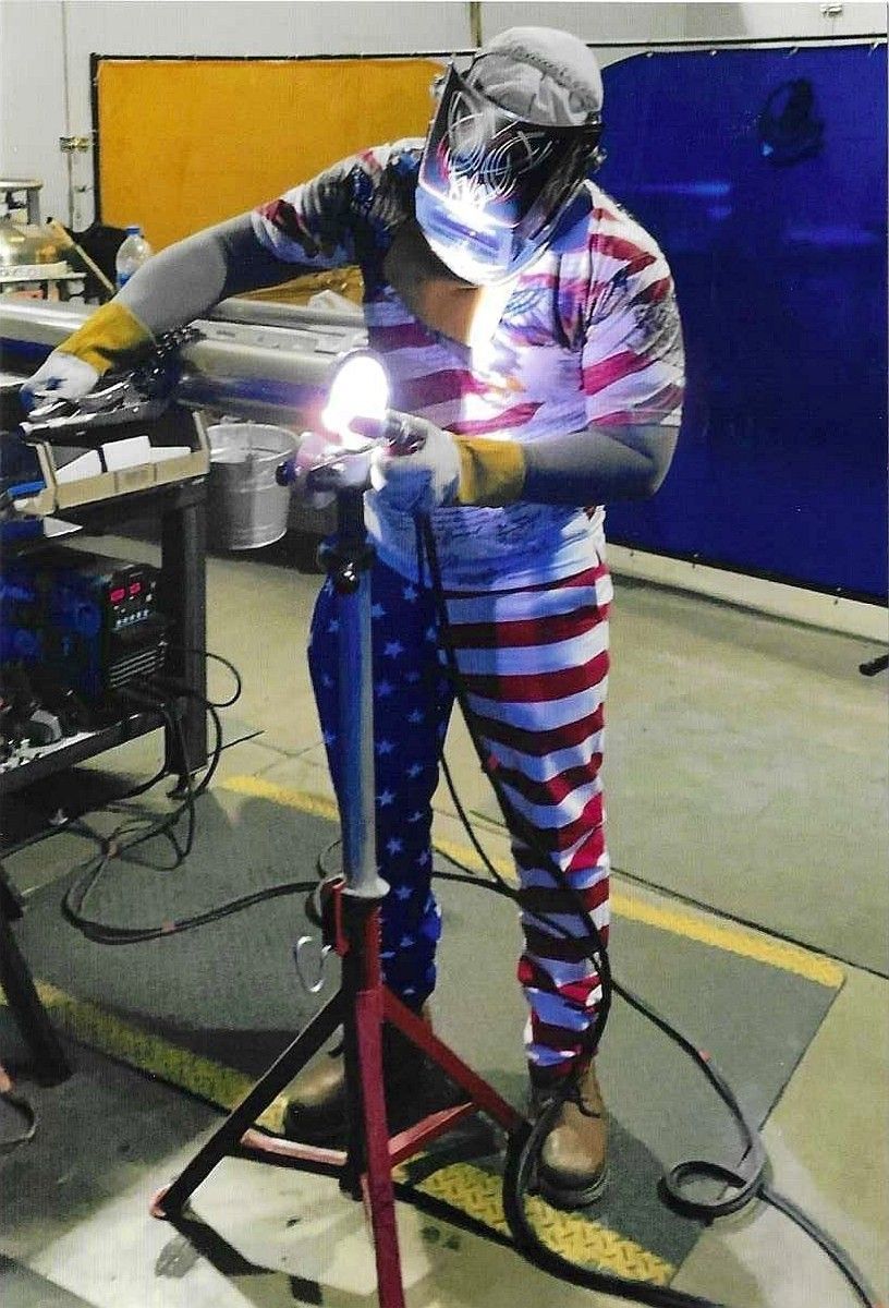 Person welding metal in a workshop, wearing a helmet and an American flag outfit.