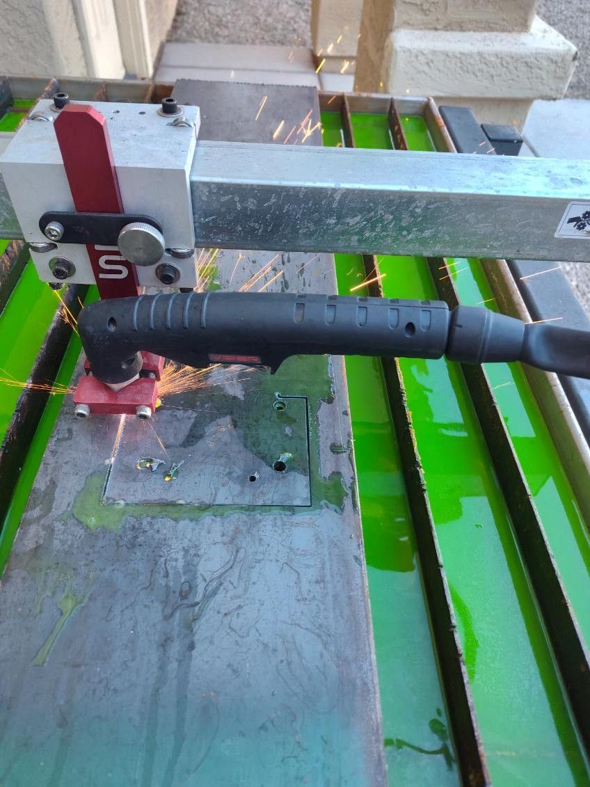 A robotic plasma cutter slices through a metal plate over a green water table, producing bright orange sparks.