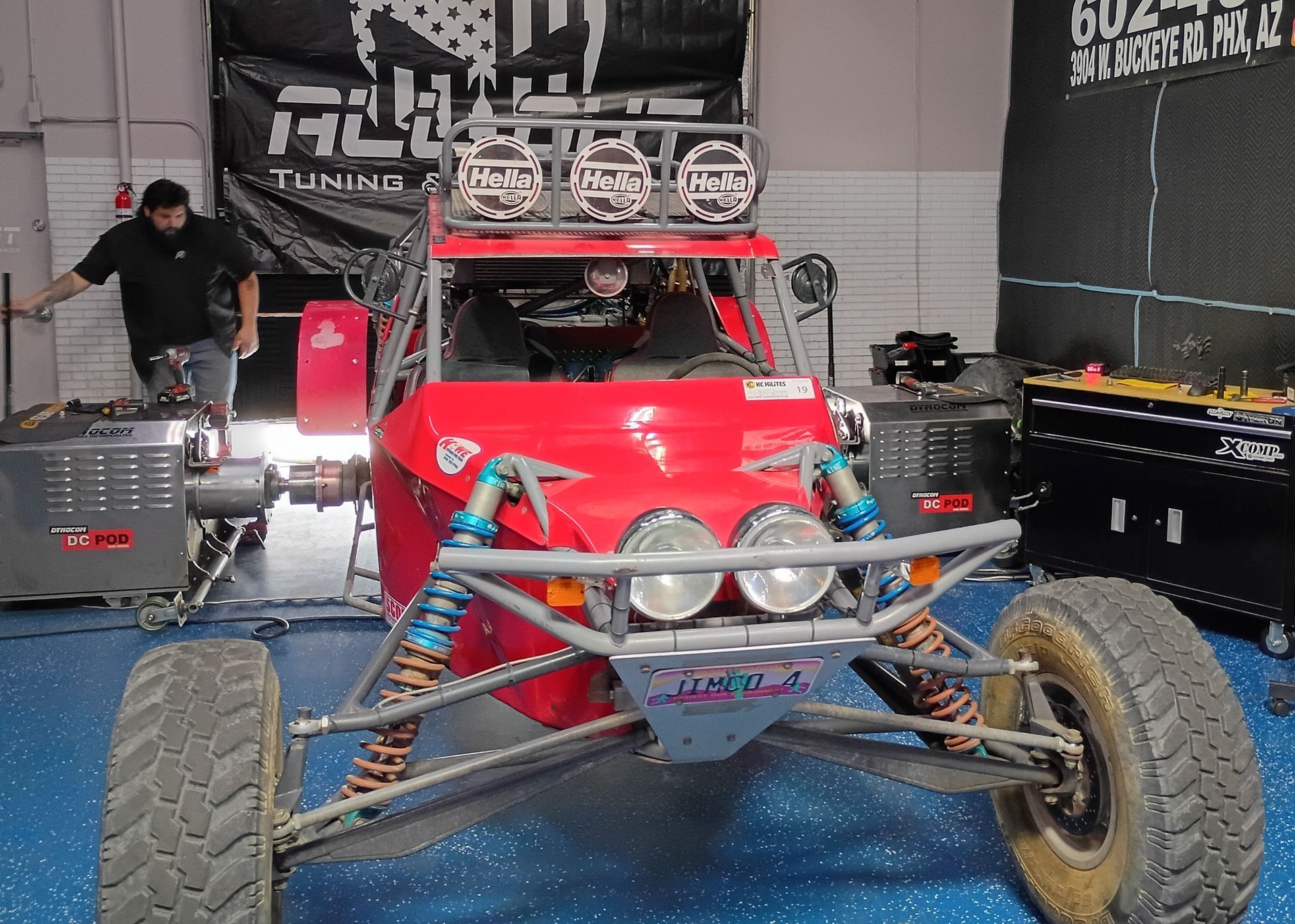 Red off-road buggy with large tires and roof gear displayed in a garage, with a person working nearby