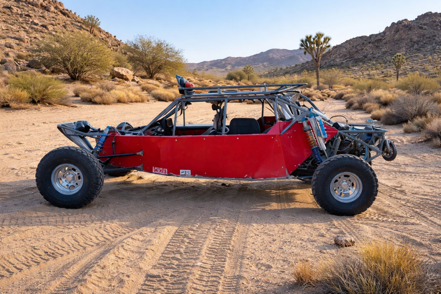 A red sand rail dune buggy parked on a sandy desert trail with hills and brush in the background.
