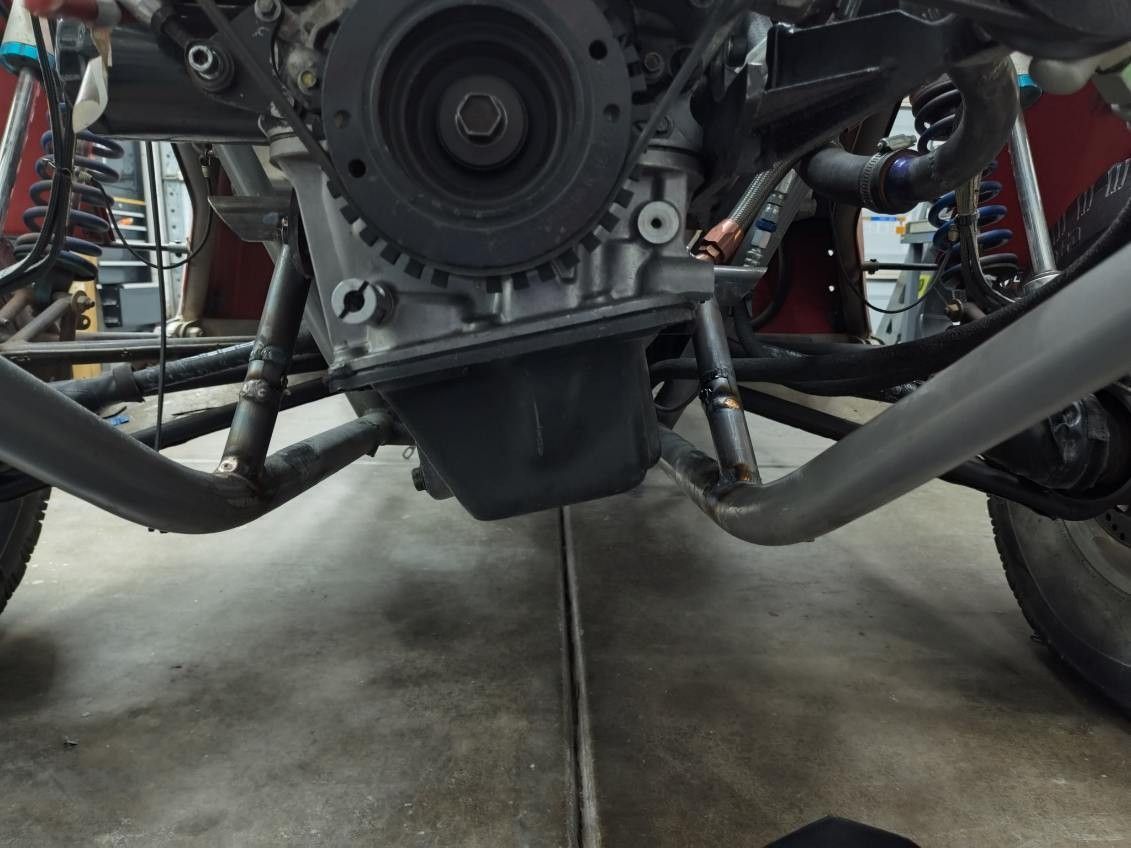 A close-up of a vehicle engine bay showing the underside of the engine, oil pan, and connected steel frame tubes.
