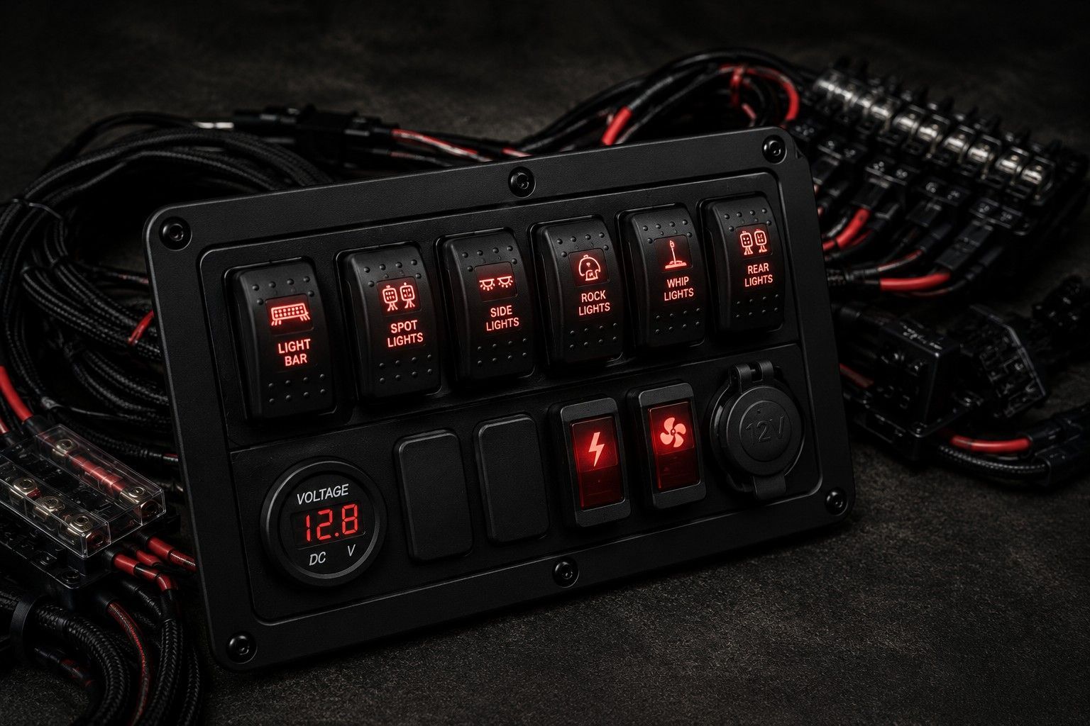 A vehicle switch panel with illuminated red labels, buttons, and toggle switches, mounted above a cluster of wiring looms.