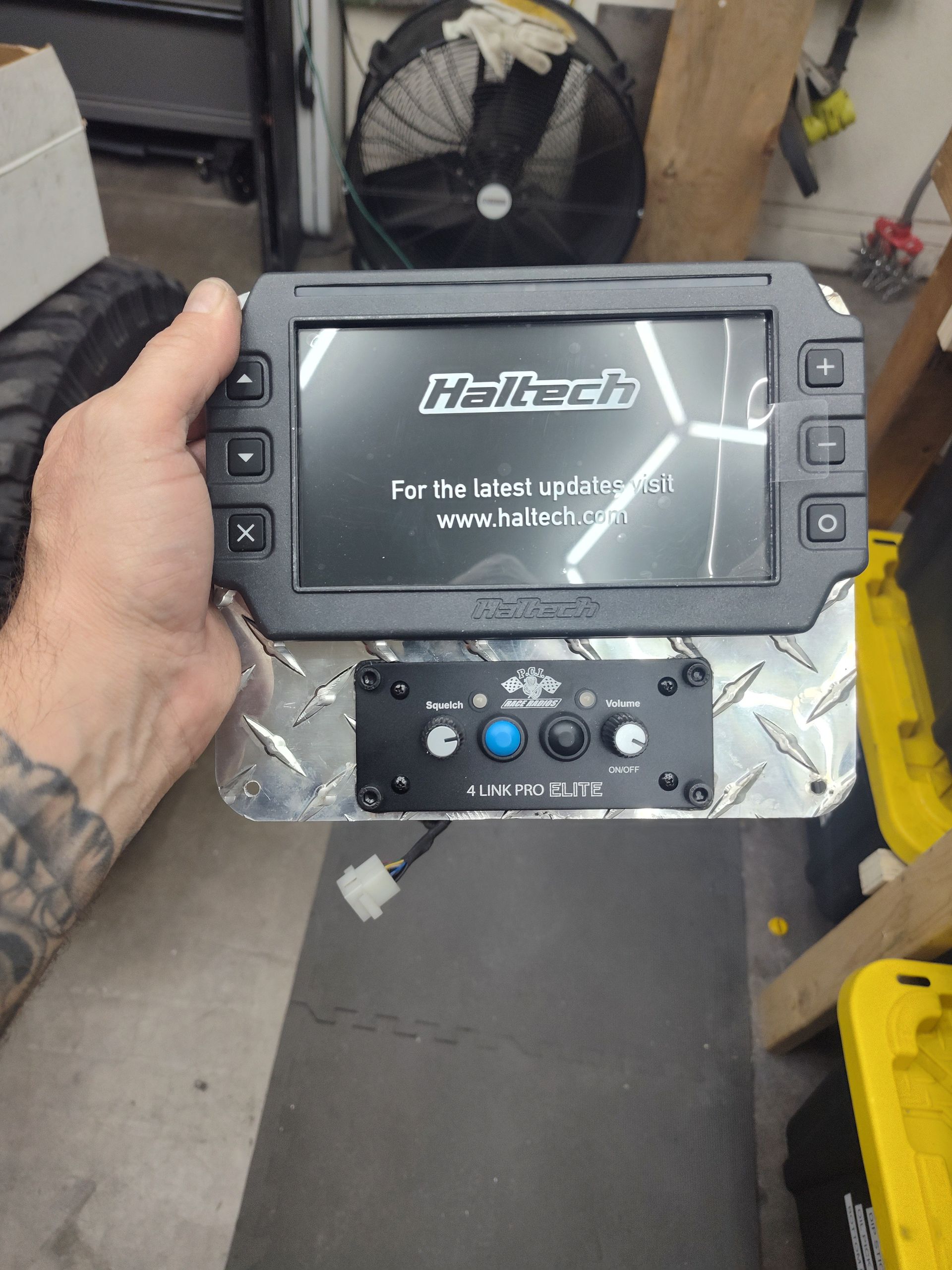 Hand holding a Haltech display and controller in a workshop, with a fan and floor mat in the background