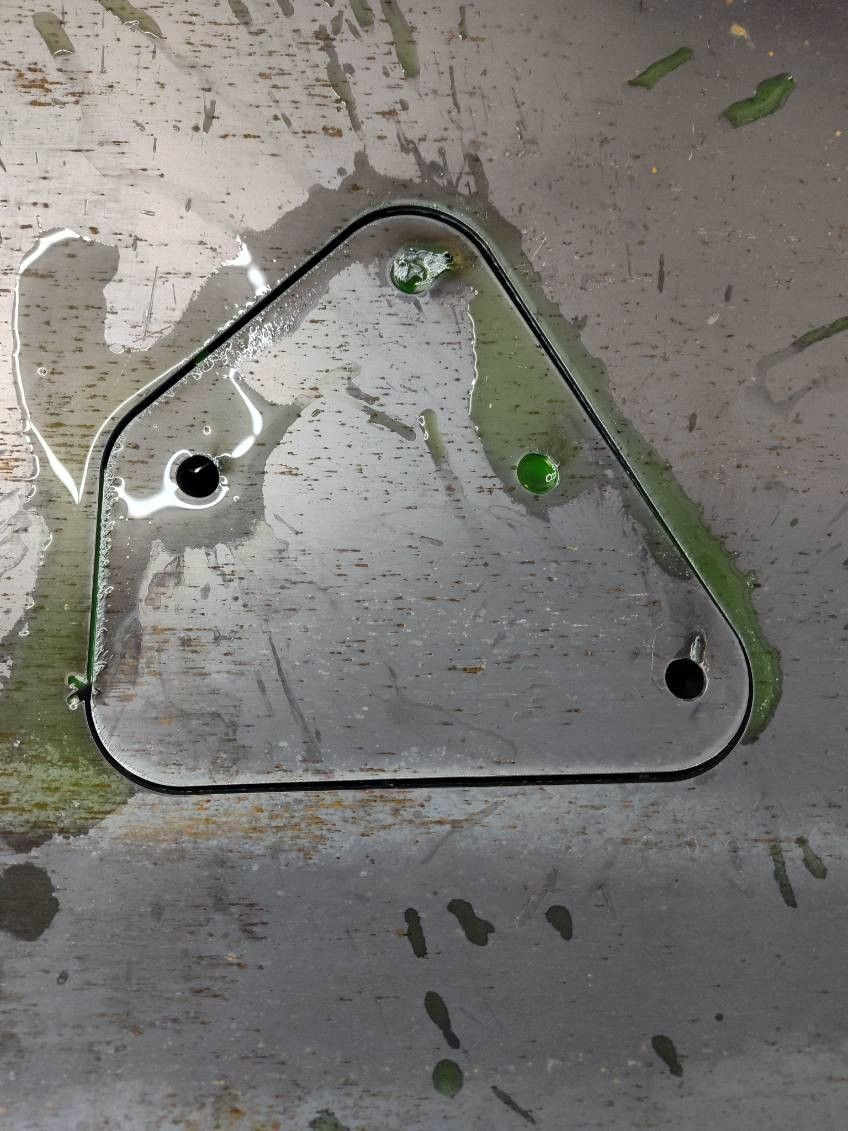 A triangular metal plate with two holes, resting on a metallic surface covered in spilled green liquid.