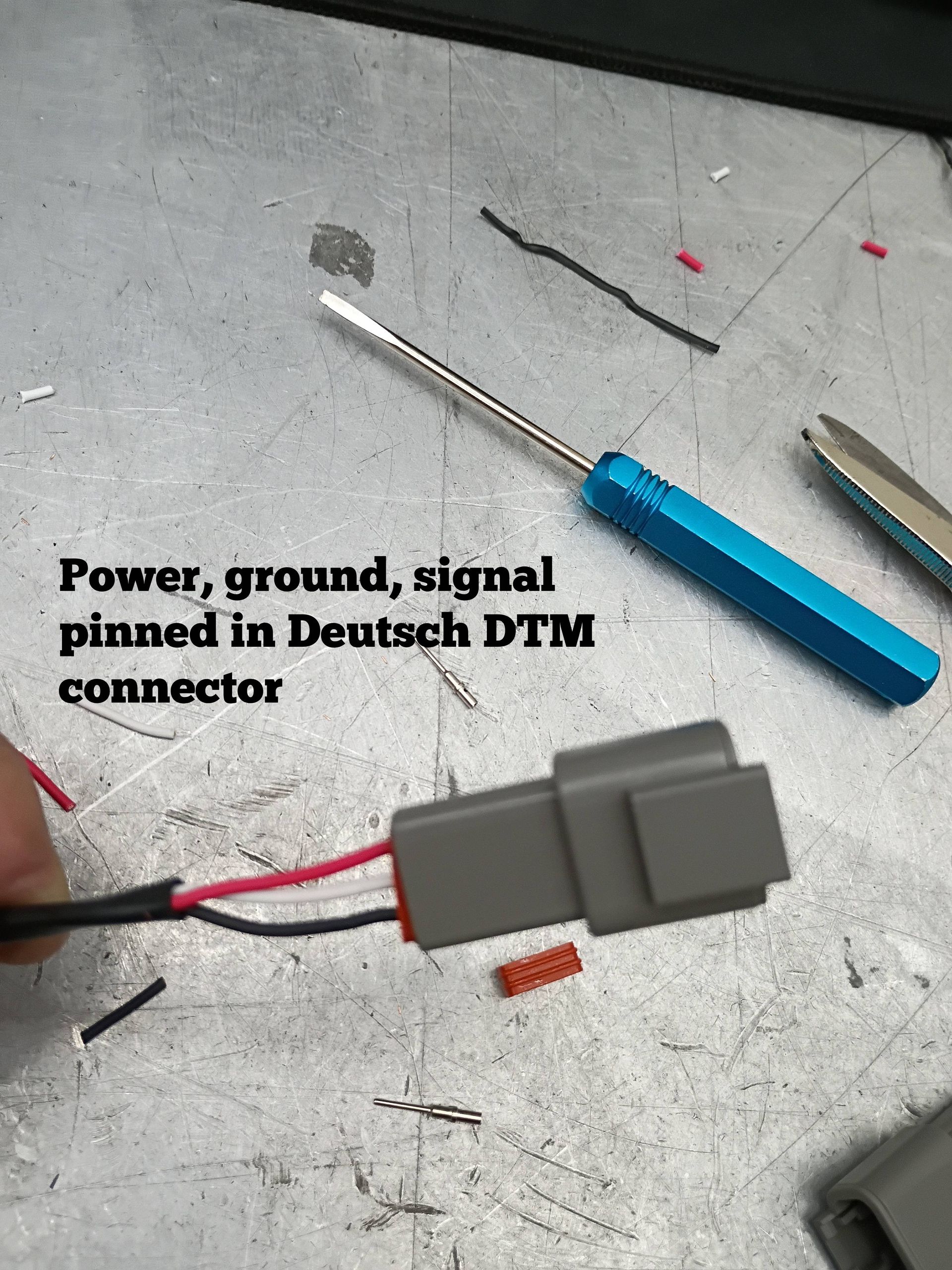 Gray Deutsch DTM connector with red and black wires on a workbench, next to a blue-handled screwdriver.