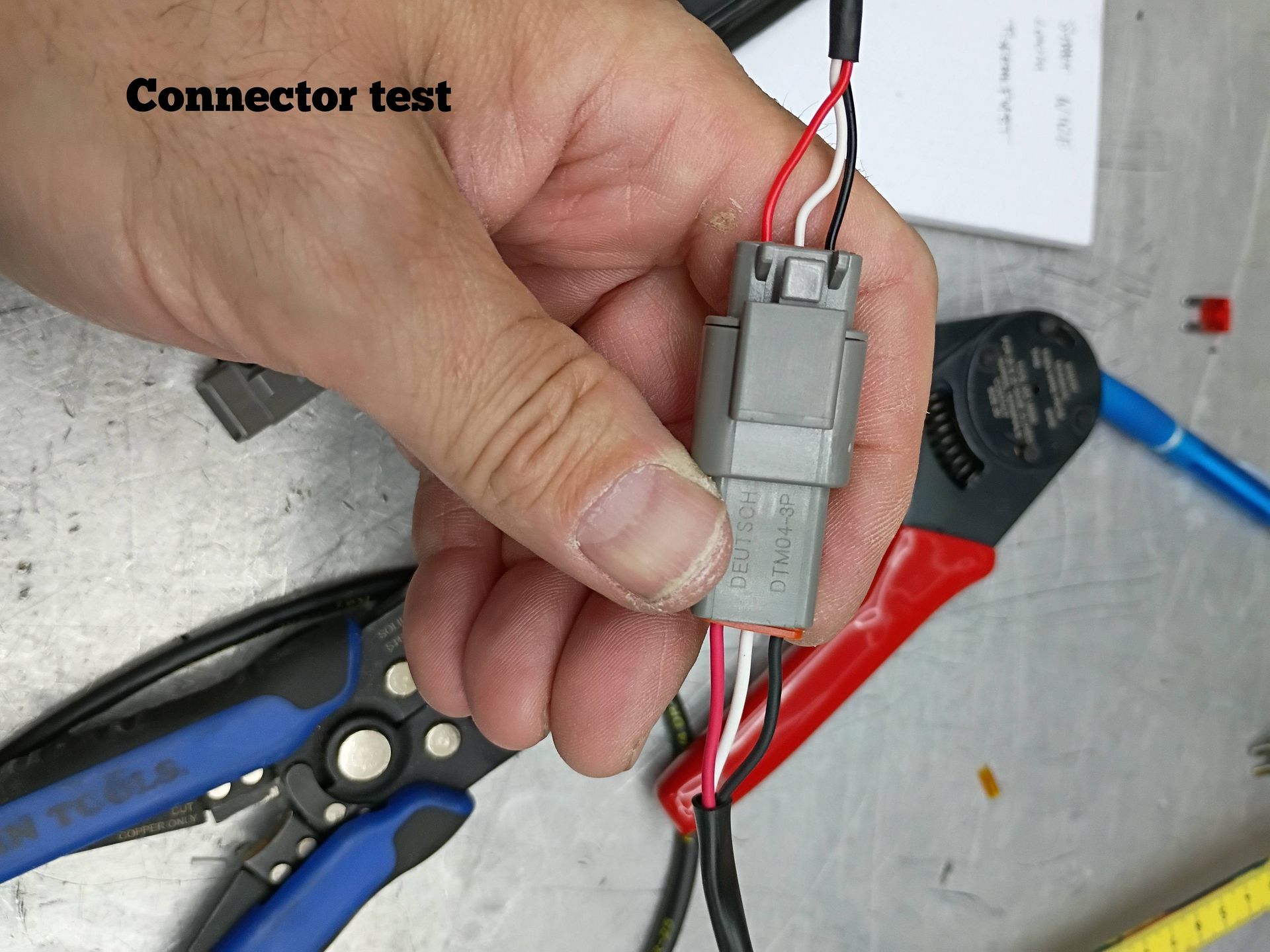 Hand holding a gray electrical connector with red and black wires on a workbench, with tools nearby