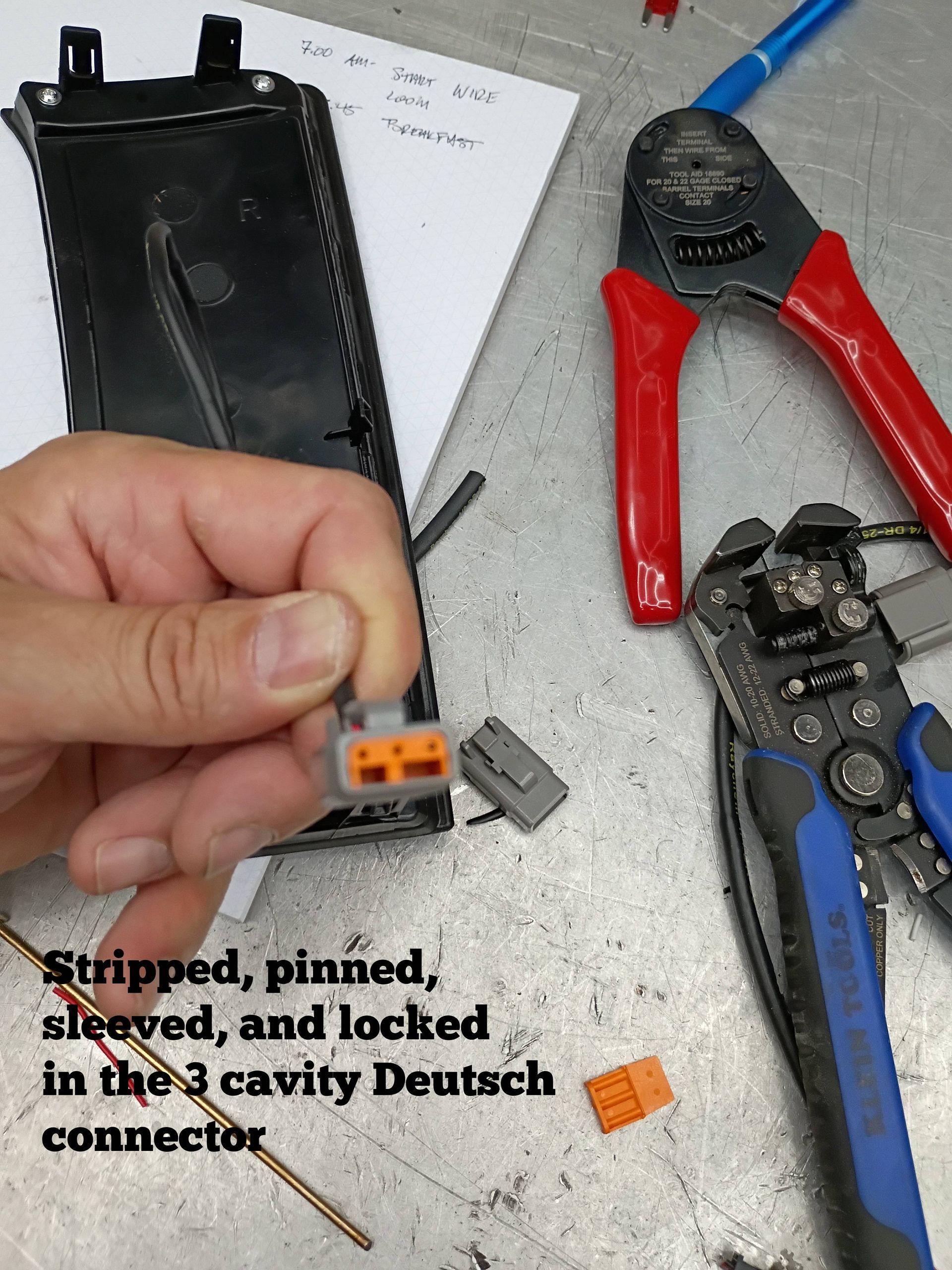 Hand holding orange Deutsch connector; pin crimper tools and stripped wire on workbench.