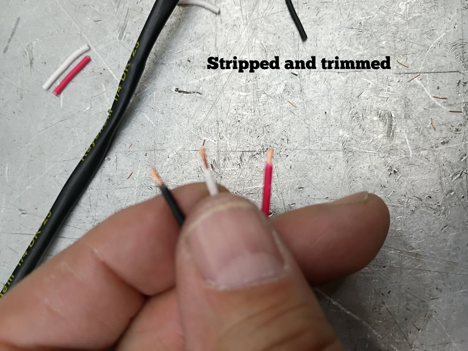 Hand holding stripped red and black wire ends on a workbench, labeled “Stripped and trimmed”