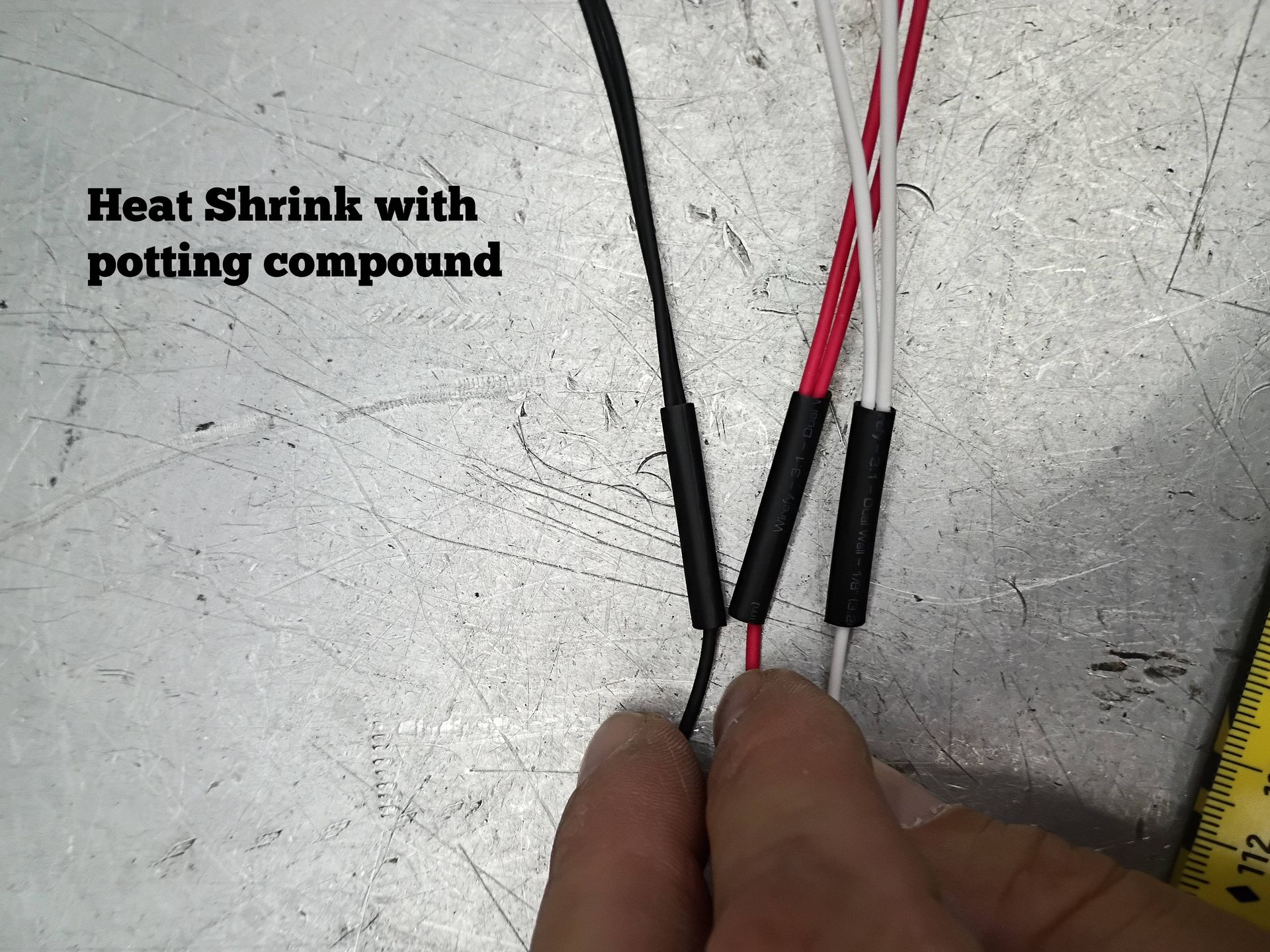 Hands holding red and black wires with heat shrink tubing and potting compound on a gray workbench