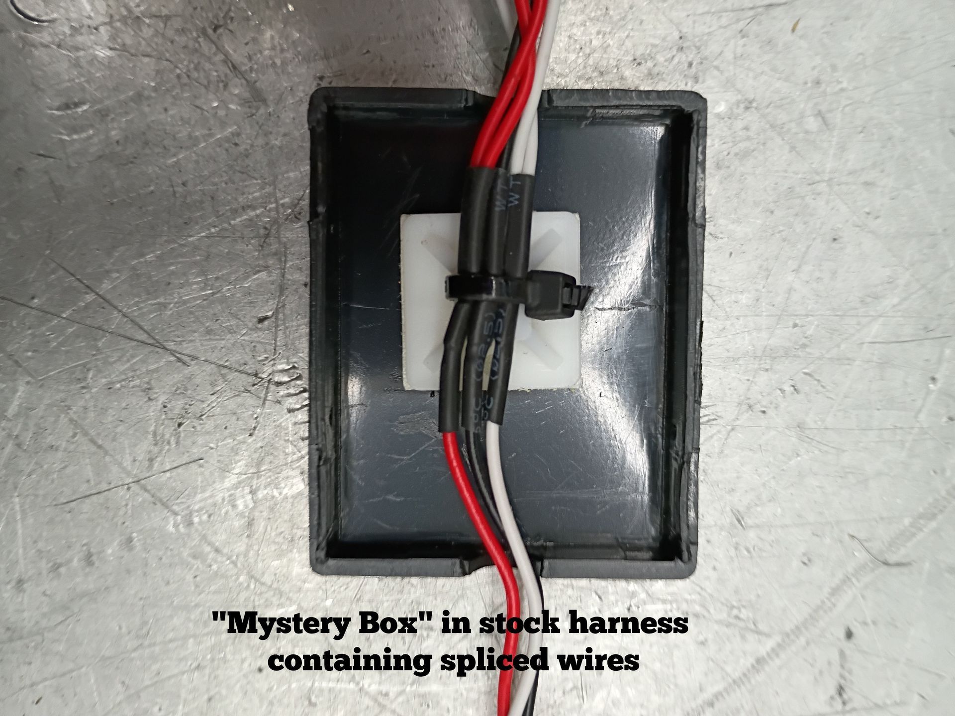 Black box in stock harness with red and black wires, secured by a zip tie on a metal surface.