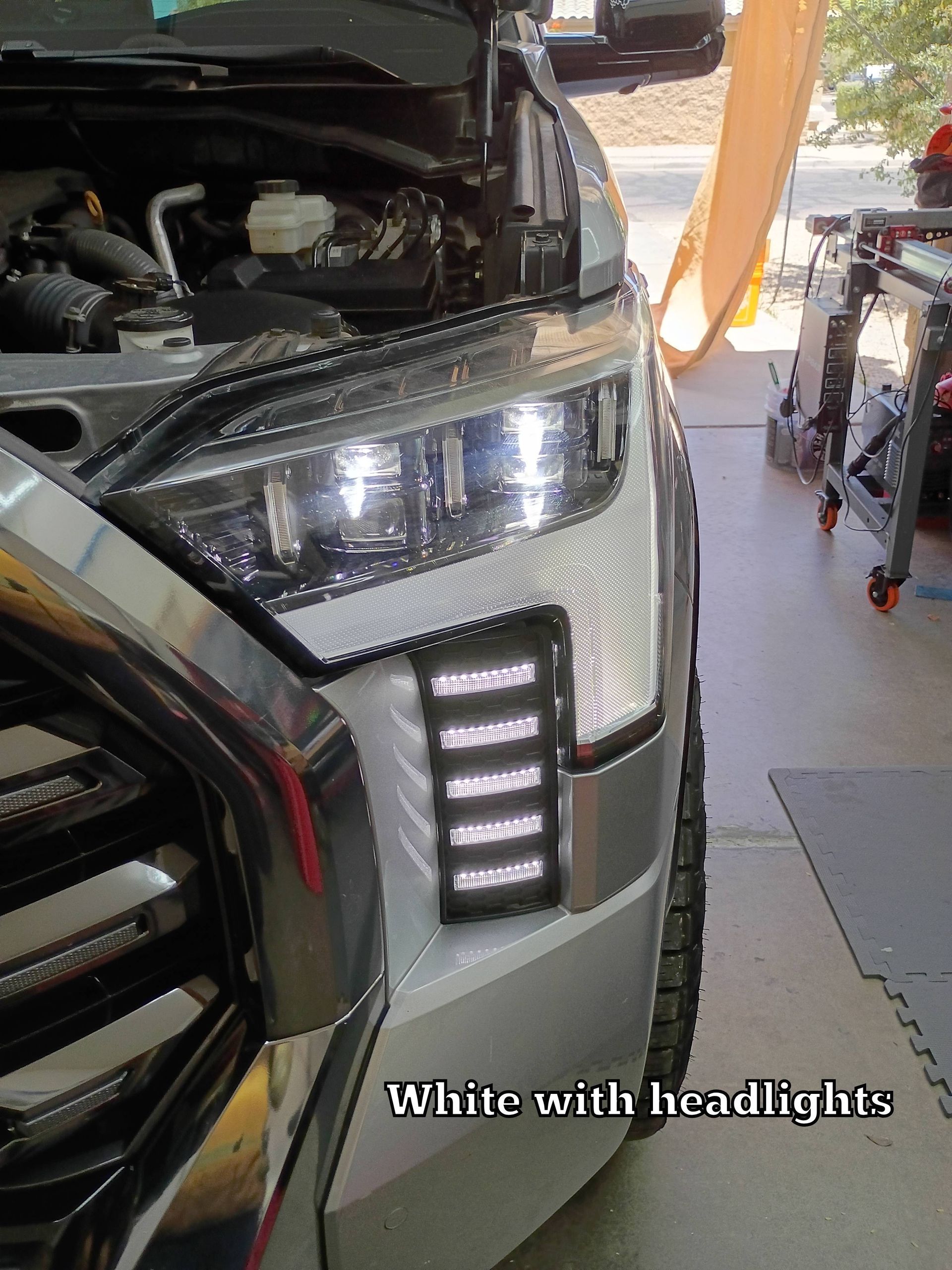 Close-up of a black car’s front headlight and open hood in a garage, with orange lift equipment nearby