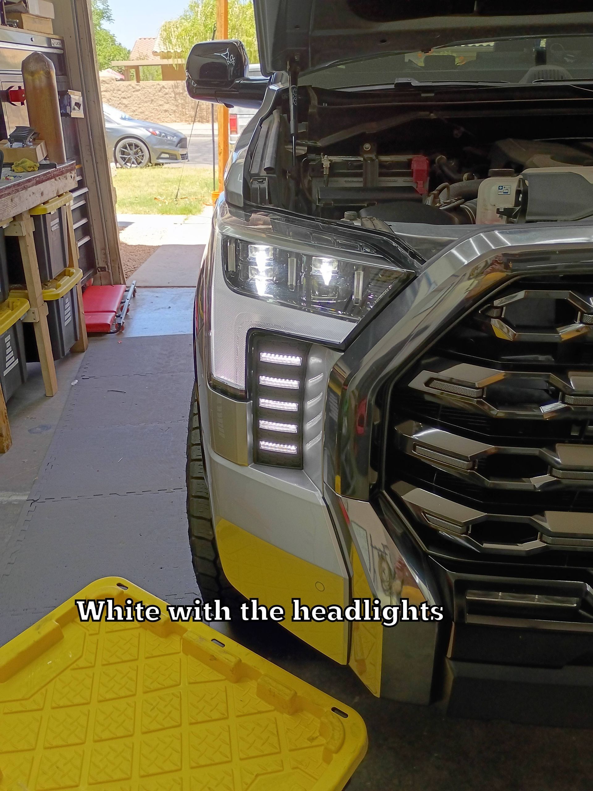 Silver SUV in a garage with hood open, showing front grille and headlights next to shelves and tools.
