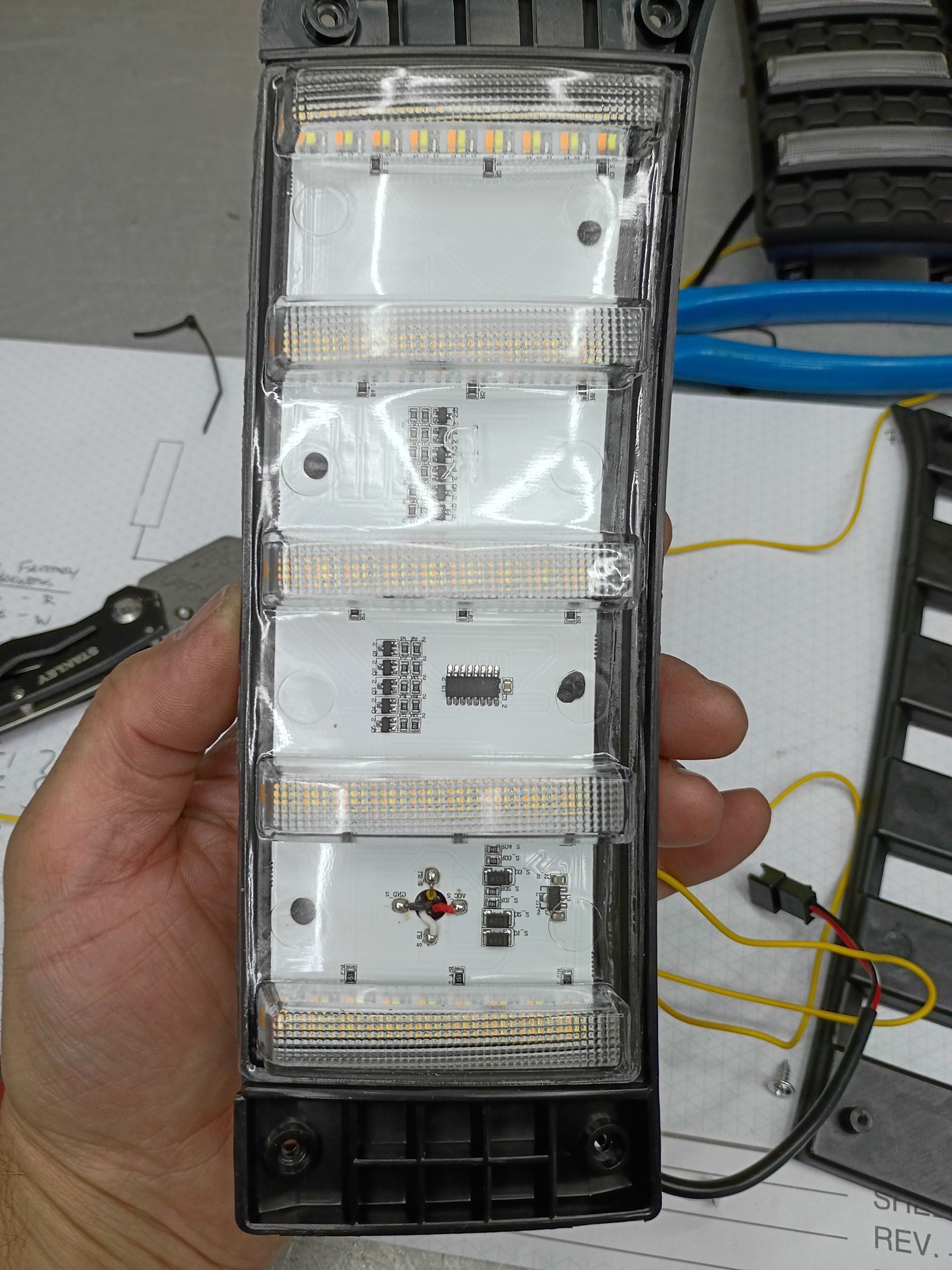 Hand holding a transparent LED light strip panel with white LEDs and black base, on a workbench.