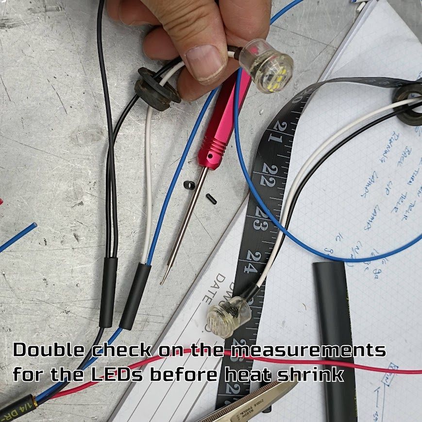 Hand holding a small LED and wiring on a workbench, with a note about checking measurements before heat shrink