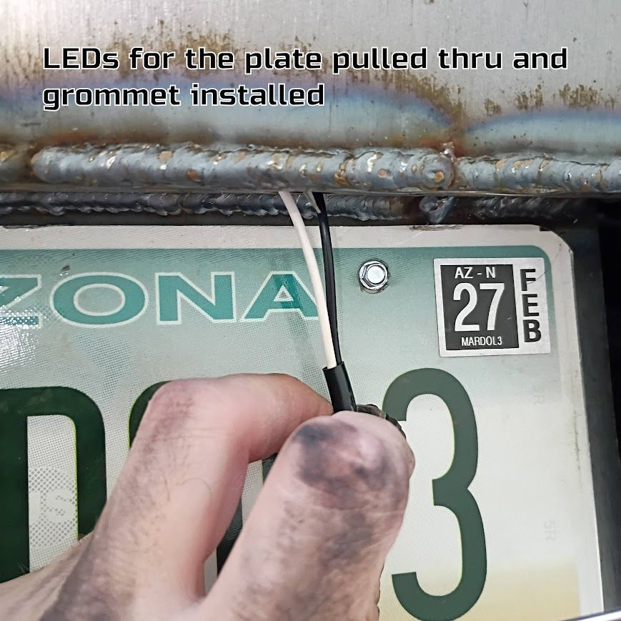 Hand installing white LED wiring through a vehicle license plate grommet on a rusty tailgate