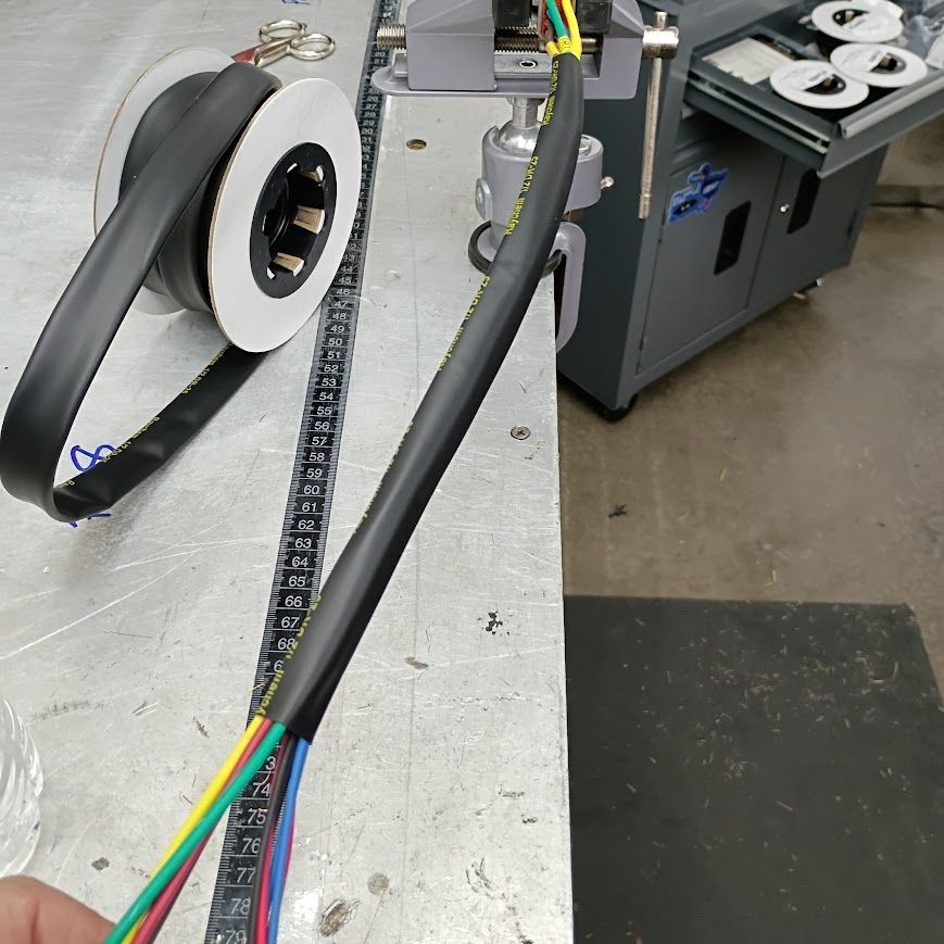 Close-up of bundled black cables with colored wires near a white spool and gray equipment on a workshop floor