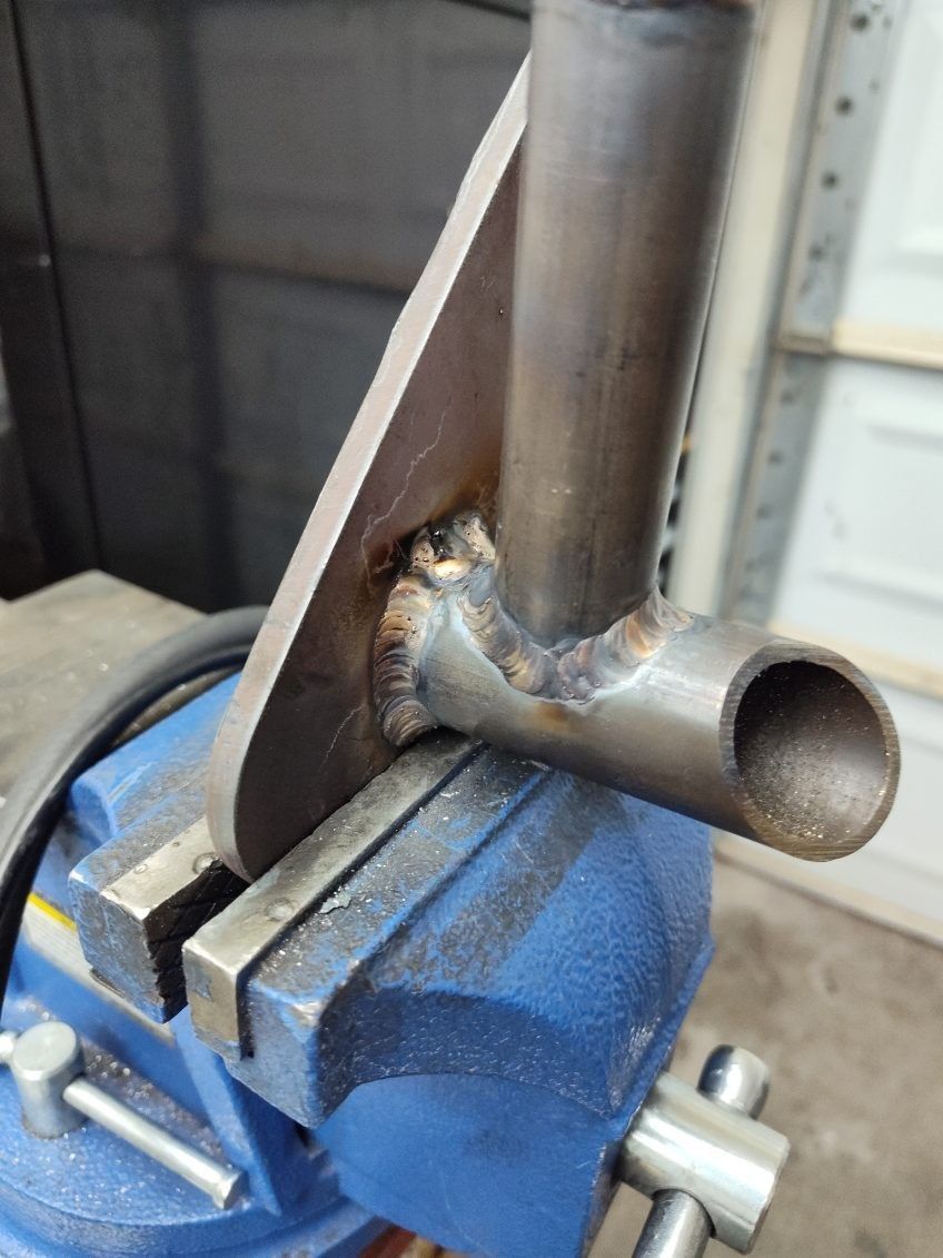 A metal T-joint assembly held in a blue bench vise, showing recent weld beads between two steel tubes and a flat plate.