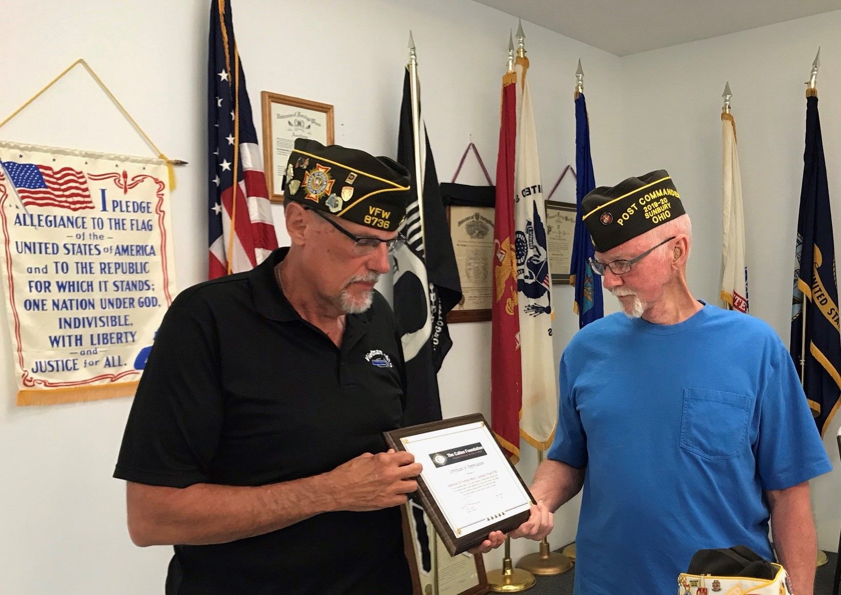 Two men are standing in front of flags and one is holding a certificate.