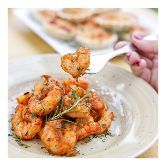 Pasta con camarones y hierbas aromáticas servida en un plato blanco, con otro plato borroso al fondo.