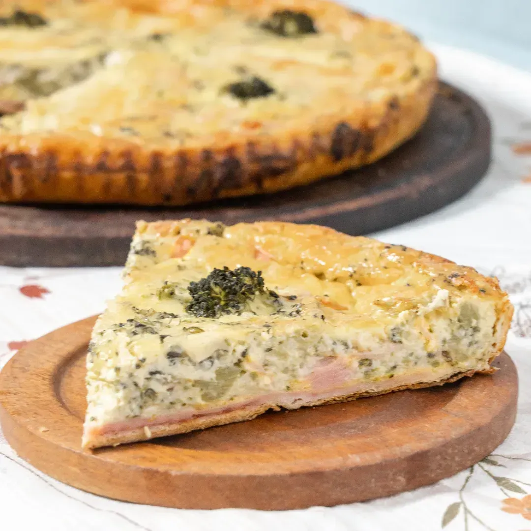 Porción de quiche salada con brócoli sobre una tabla de madera, con la quiche entera desenfocada al fondo.