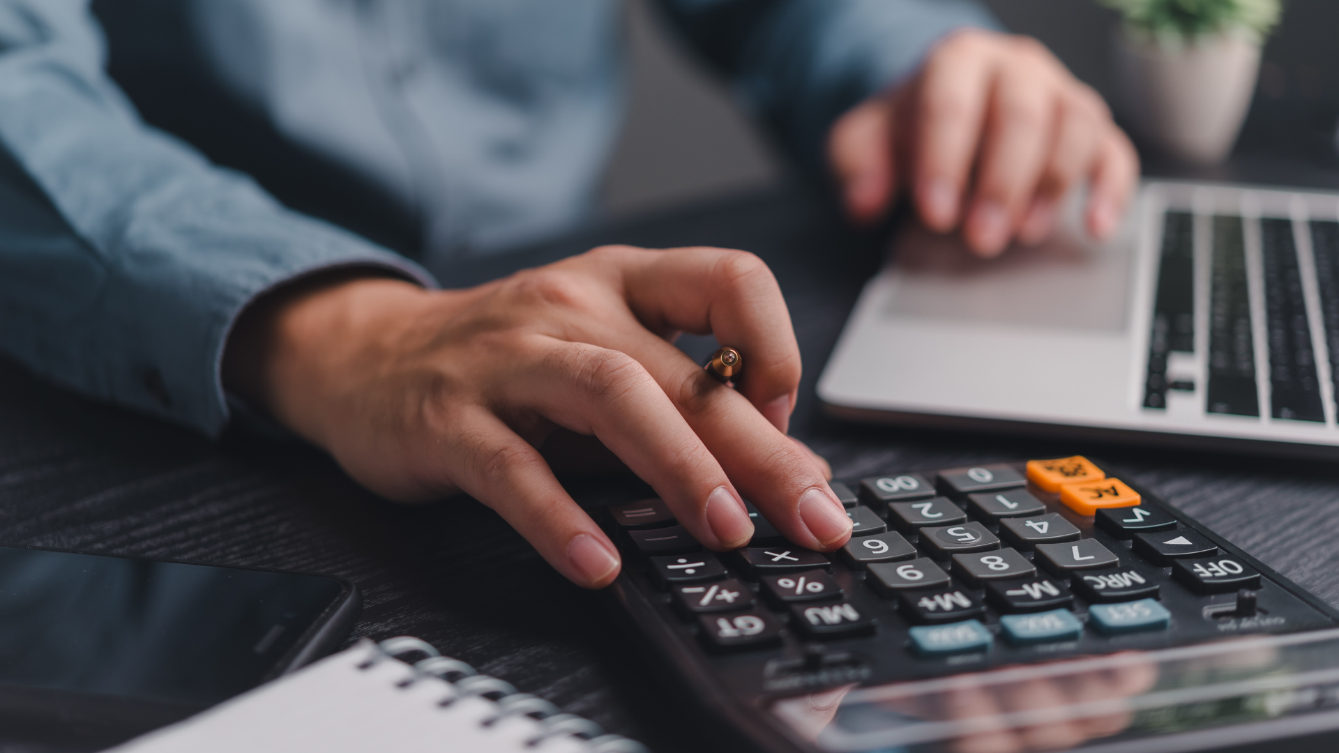 Hands using a calculator beside a laptop on a desk with paperwork