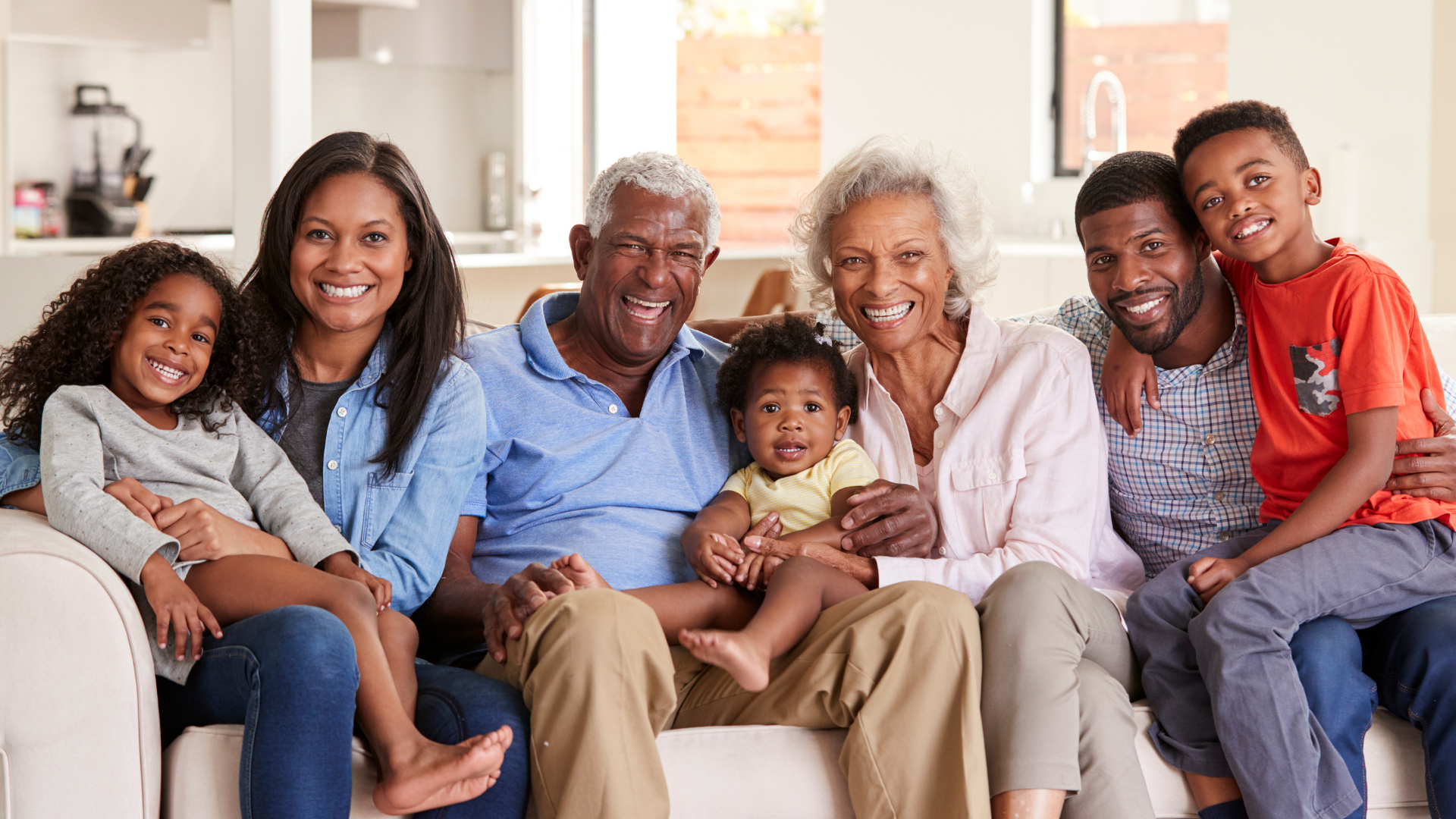 Multi-generational family sitting together on a couch, smiling in a bright living room.