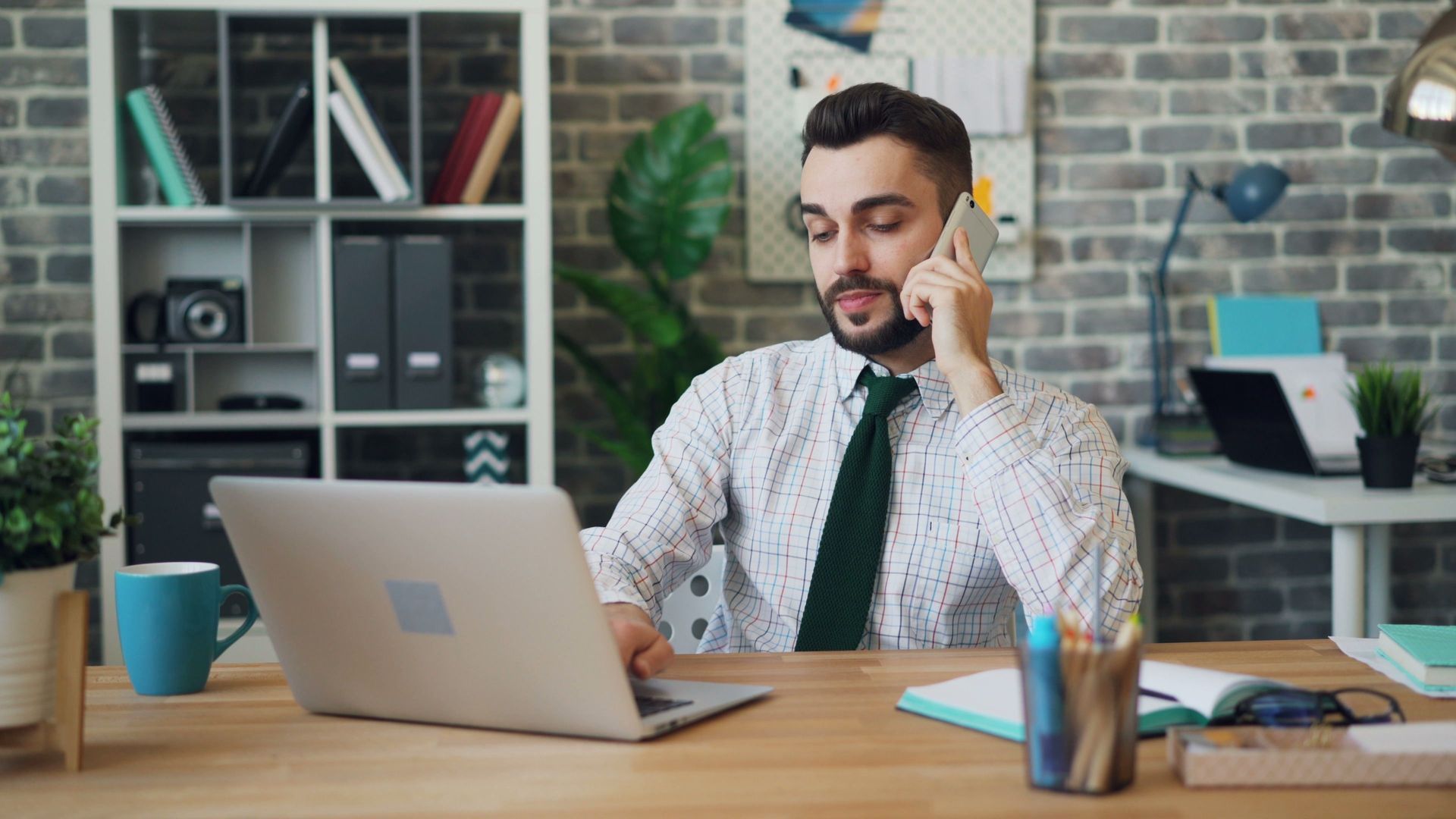 Man on a phone call beside a laptop at a desk in a bright office